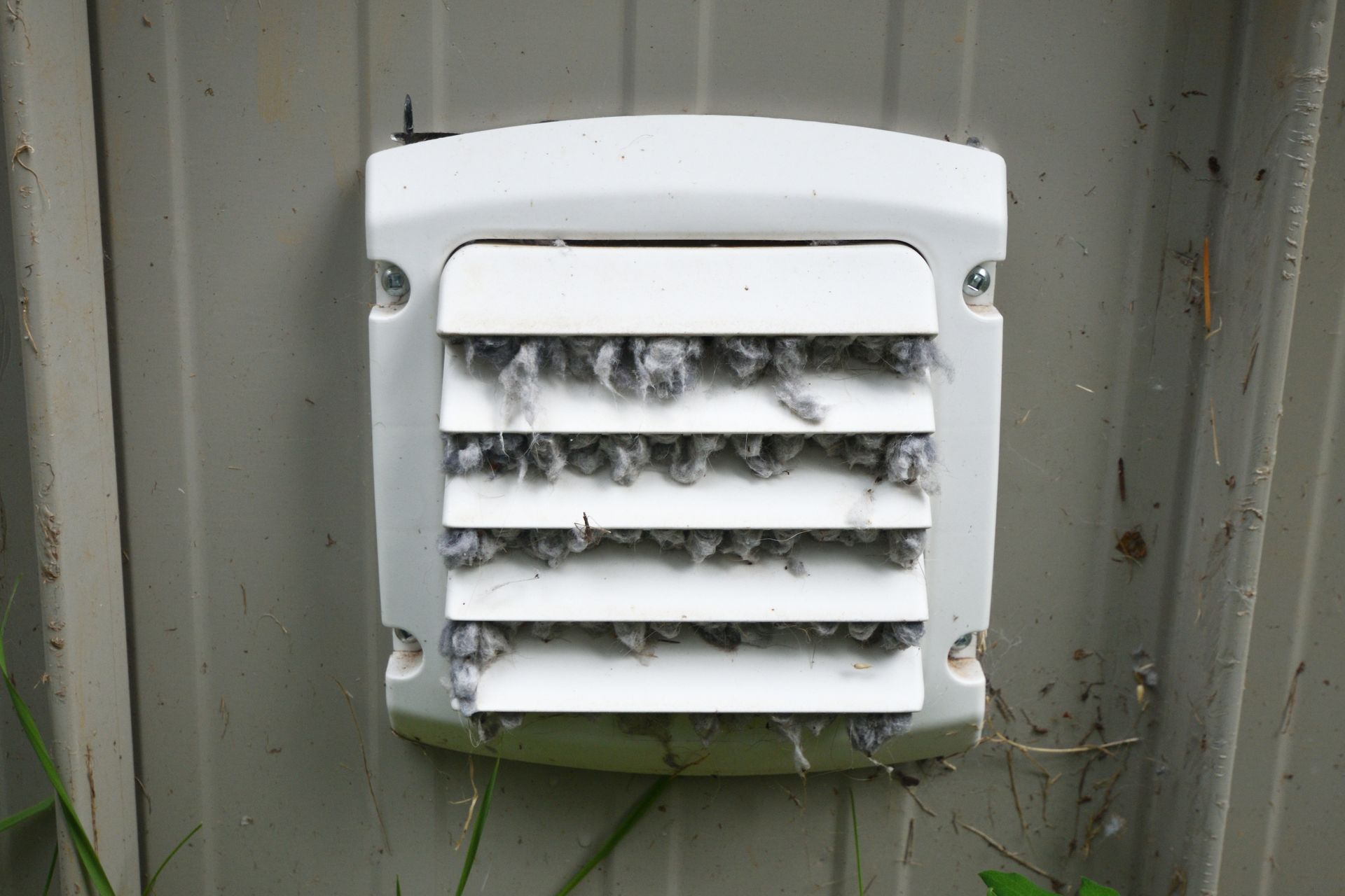 White vent cover on a gray wall, with dirt and cobwebs caught in the slats.