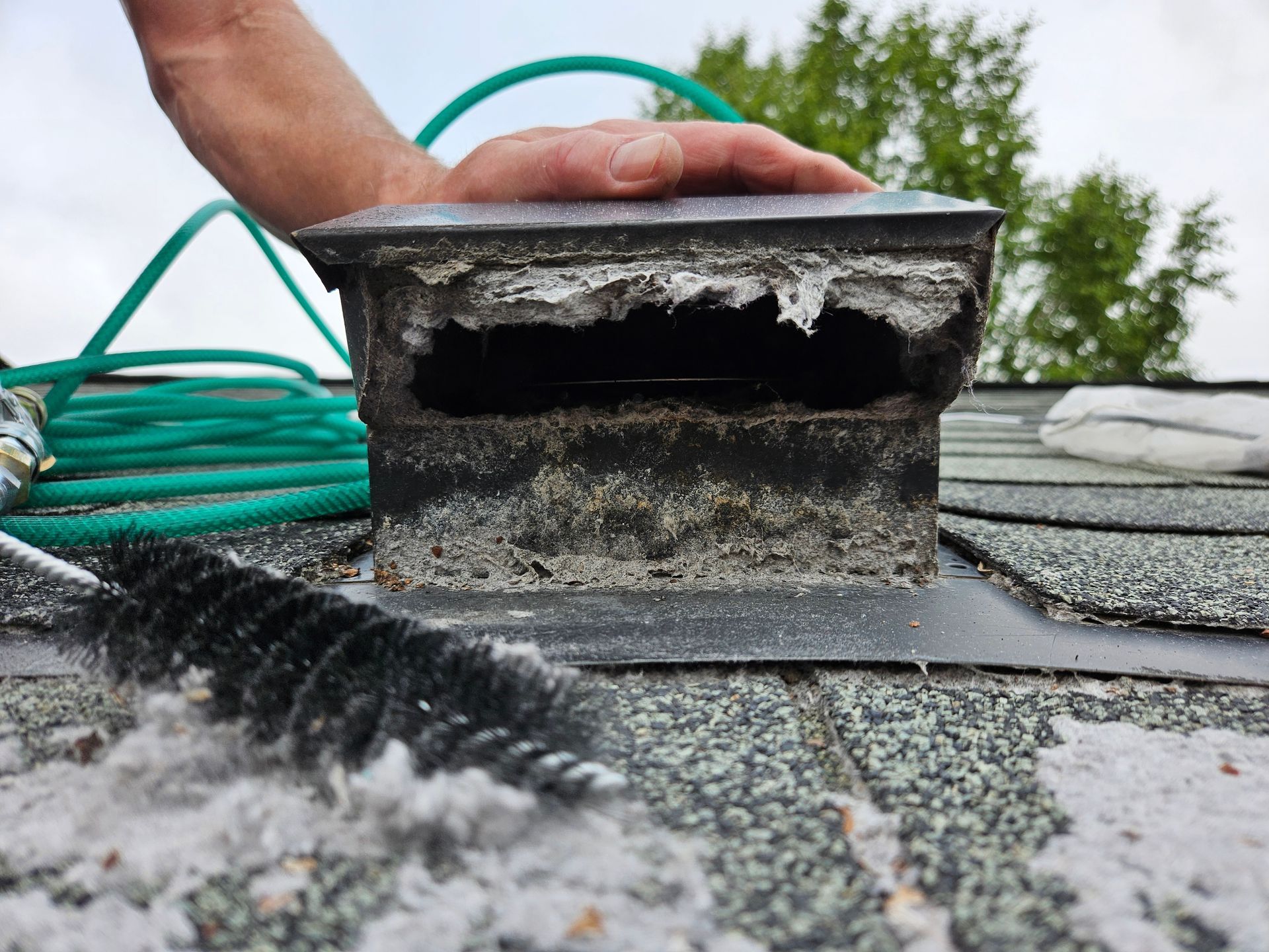Damaged roof vent with charred debris and a hand nearby on a shingled roof
