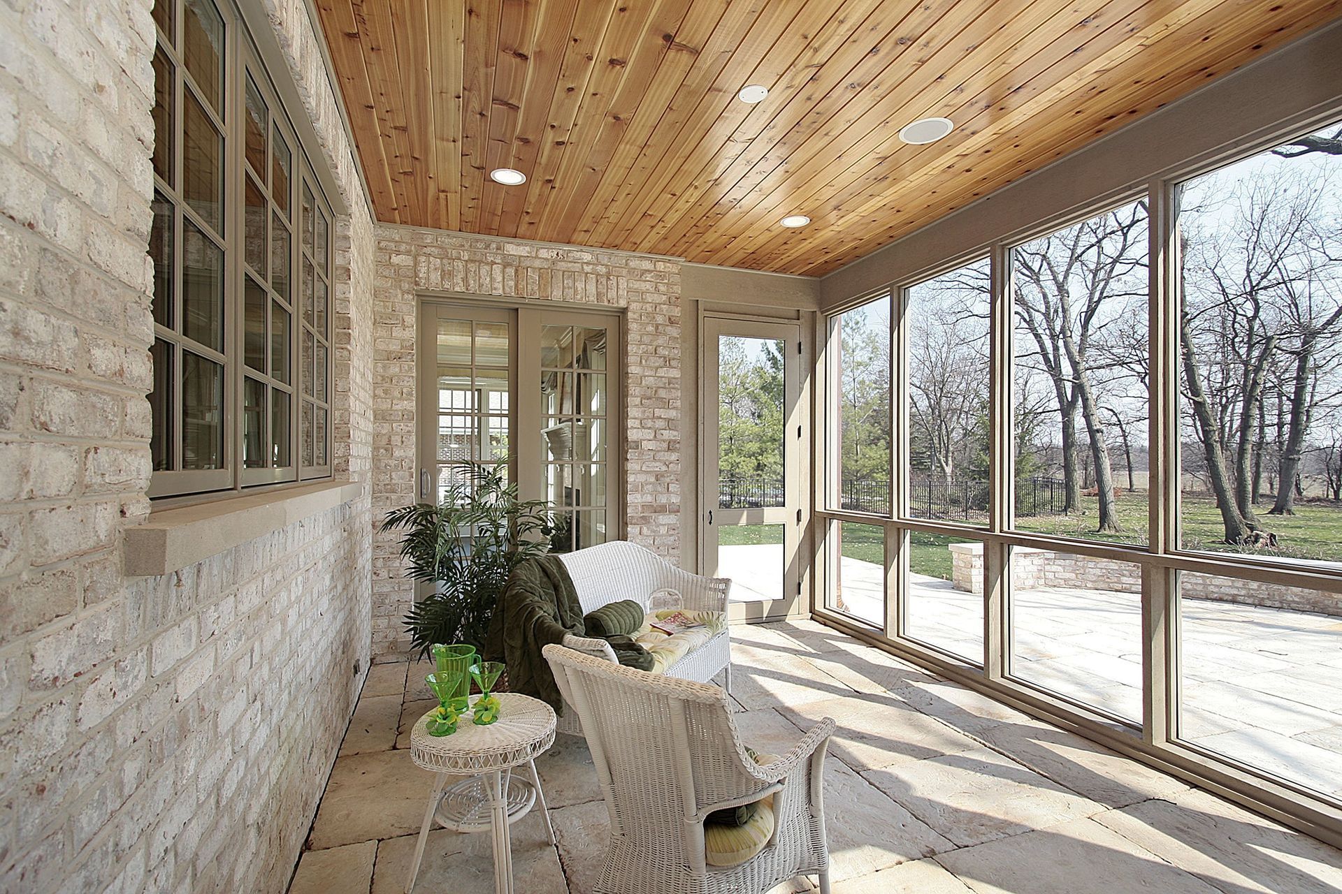 Sunroom with wicker chairs, brick wall, wood ceiling, and floor-to-ceiling windows overlooking bare trees