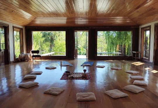 En un estudio de yoga, una persona se arrodilla junto a un alumno que está sobre un cojín en el suelo.