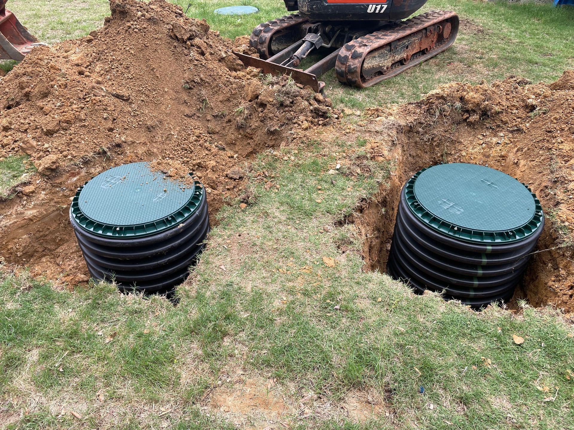 A small excavator is digging a hole in the ground next to two septic tanks.