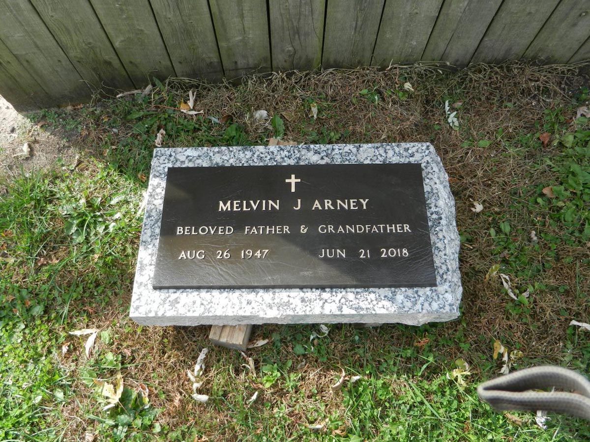 A gravestone for melvin arney is sitting in the grass next to a wooden fence