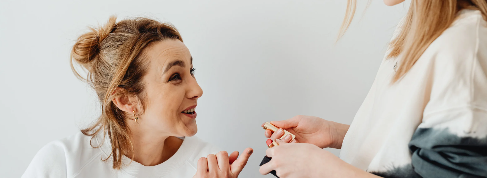Woman with bun talking about beauty treatments to person holding lipstick. Bright background.