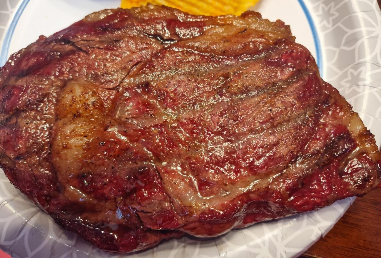 Grilled ribeye steak, medium-rare, on a paper plate. The steak is reddish-brown with visible grill marks and fat marbling.