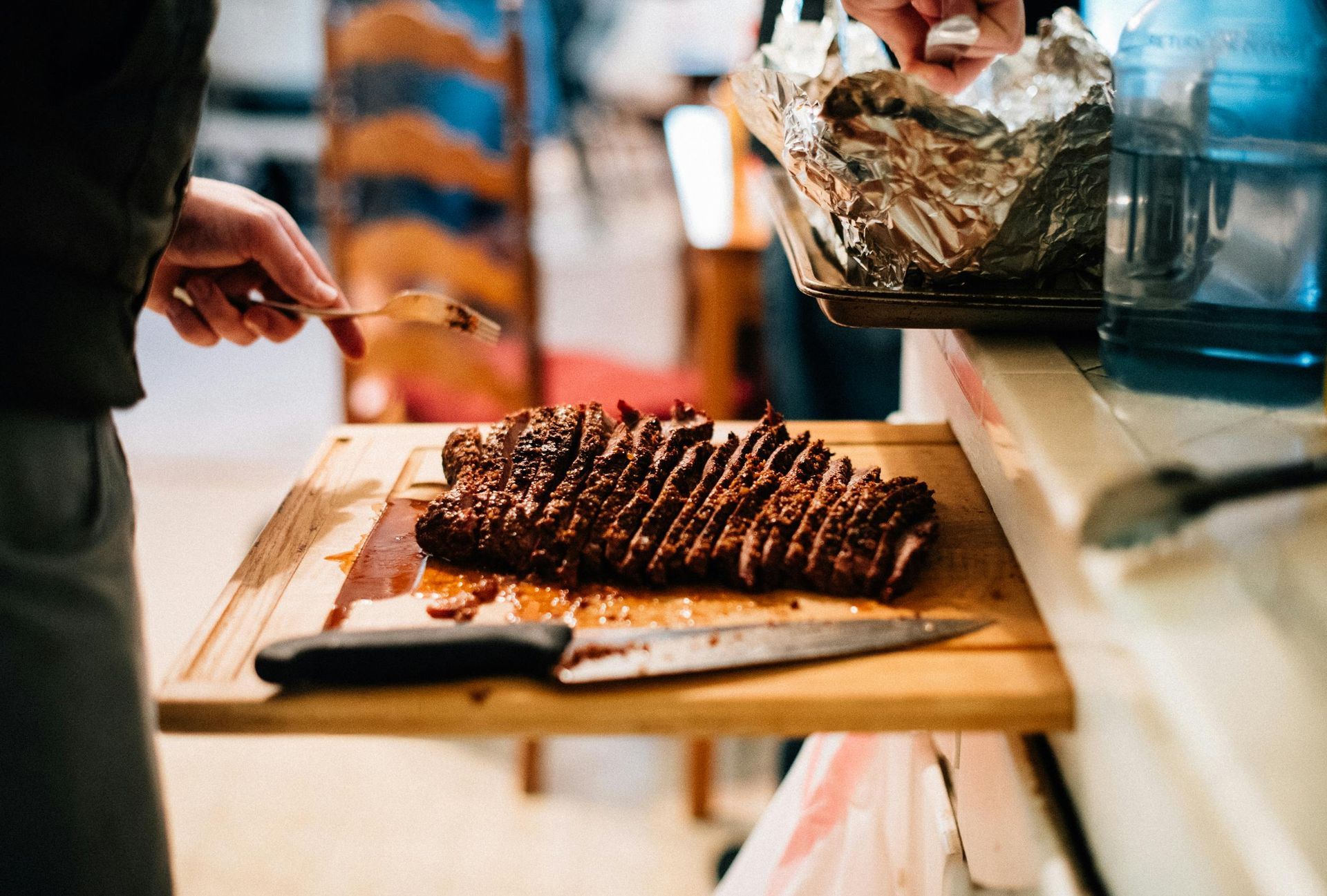 Person slicing a cooked, seasoned meat loaf on a wooden cutting board. Another person holds a pan covered in foil, with a water jug visible.