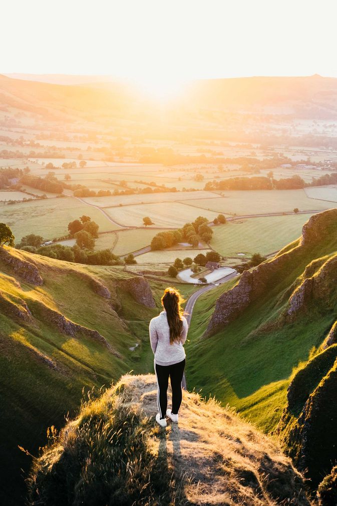 A woman is standing on top of a hill overlooking a valley at sunset.