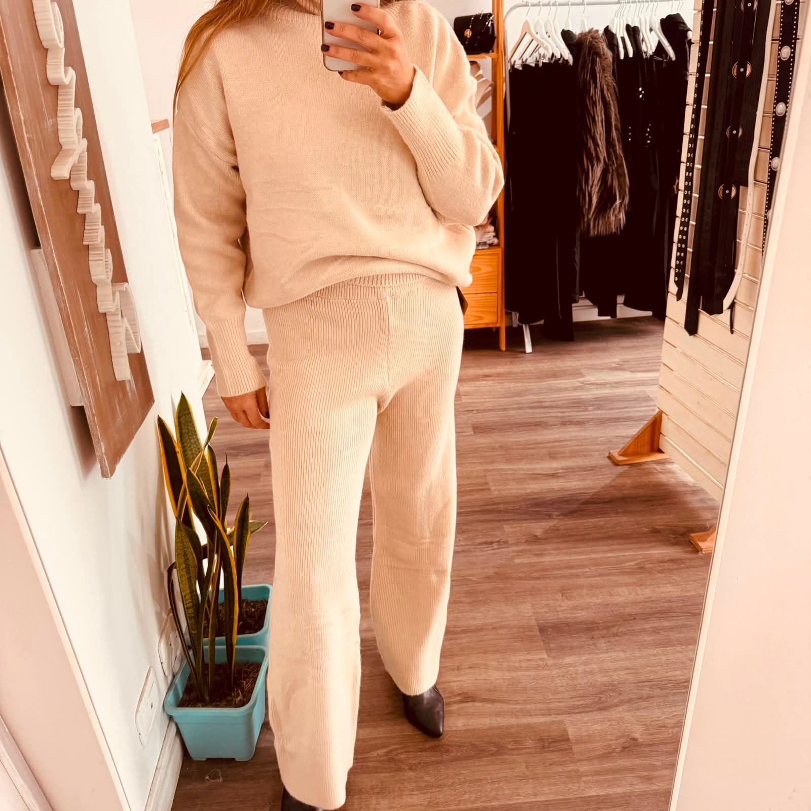 Persona de pie en el interior de una vivienda, vestida con pantalones y camiseta de punto marrón.