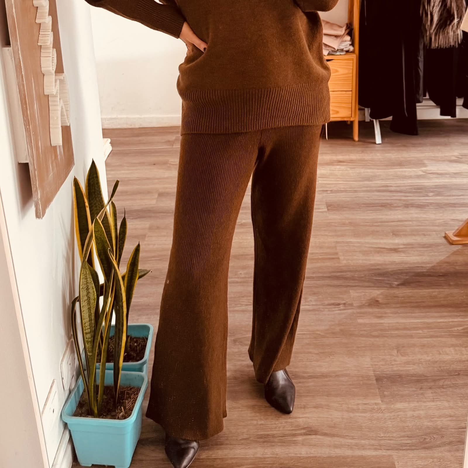 Persona de pie en el interior de una vivienda, vestida con pantalones y camiseta de punto marrón.