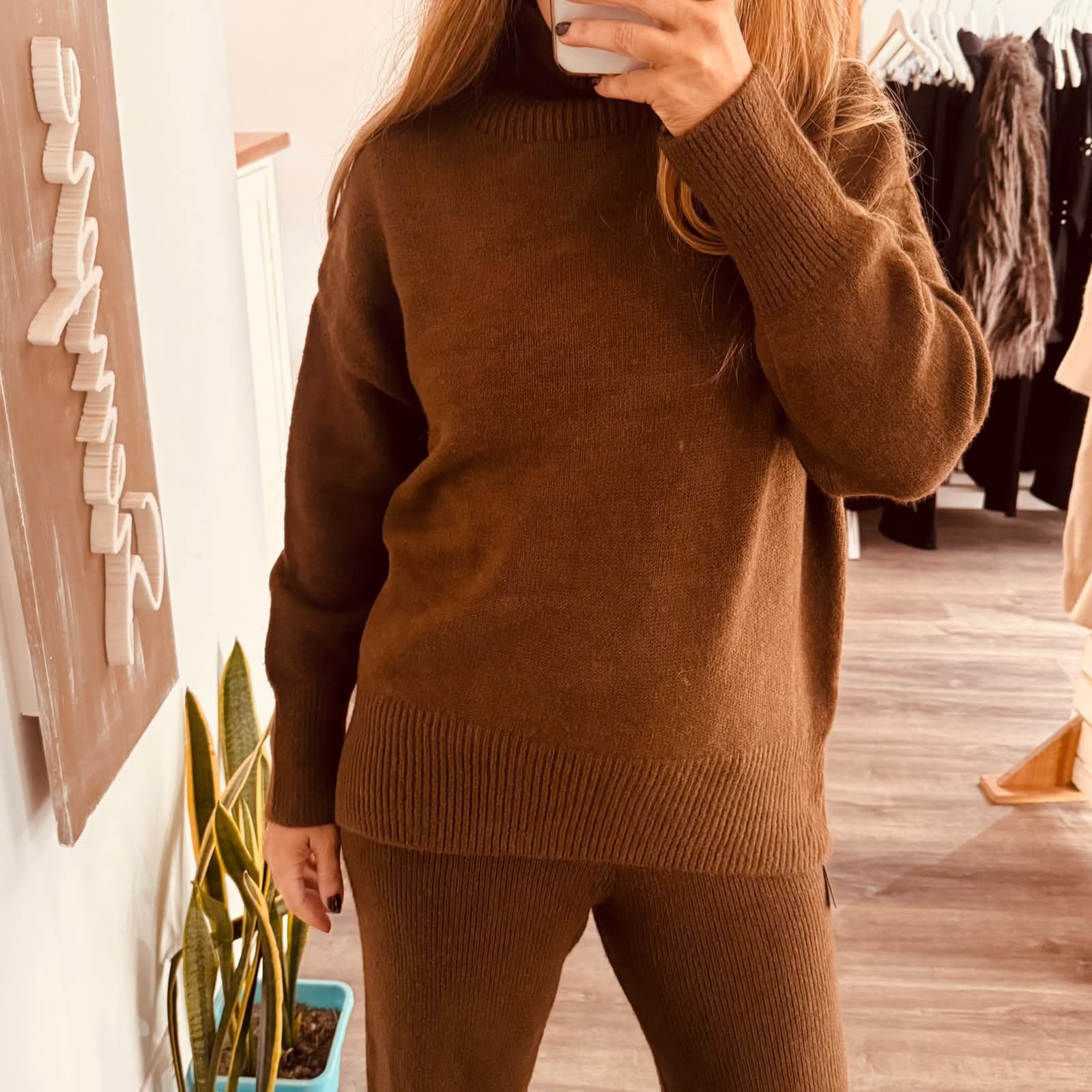 Persona con suéter marrón y pantalones de punto tomándose una selfie frente al espejo en el interior de una casa.