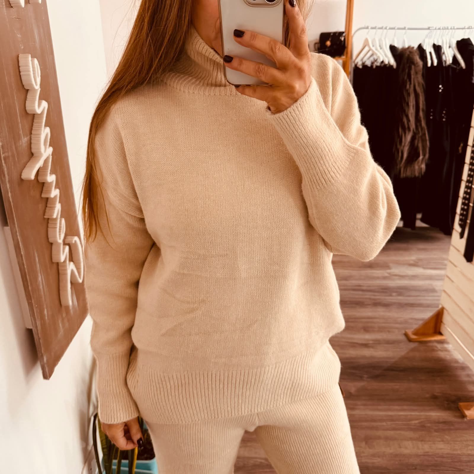 Persona con suéter marrón y pantalones de punto tomándose una selfie frente al espejo en el interior de una casa.