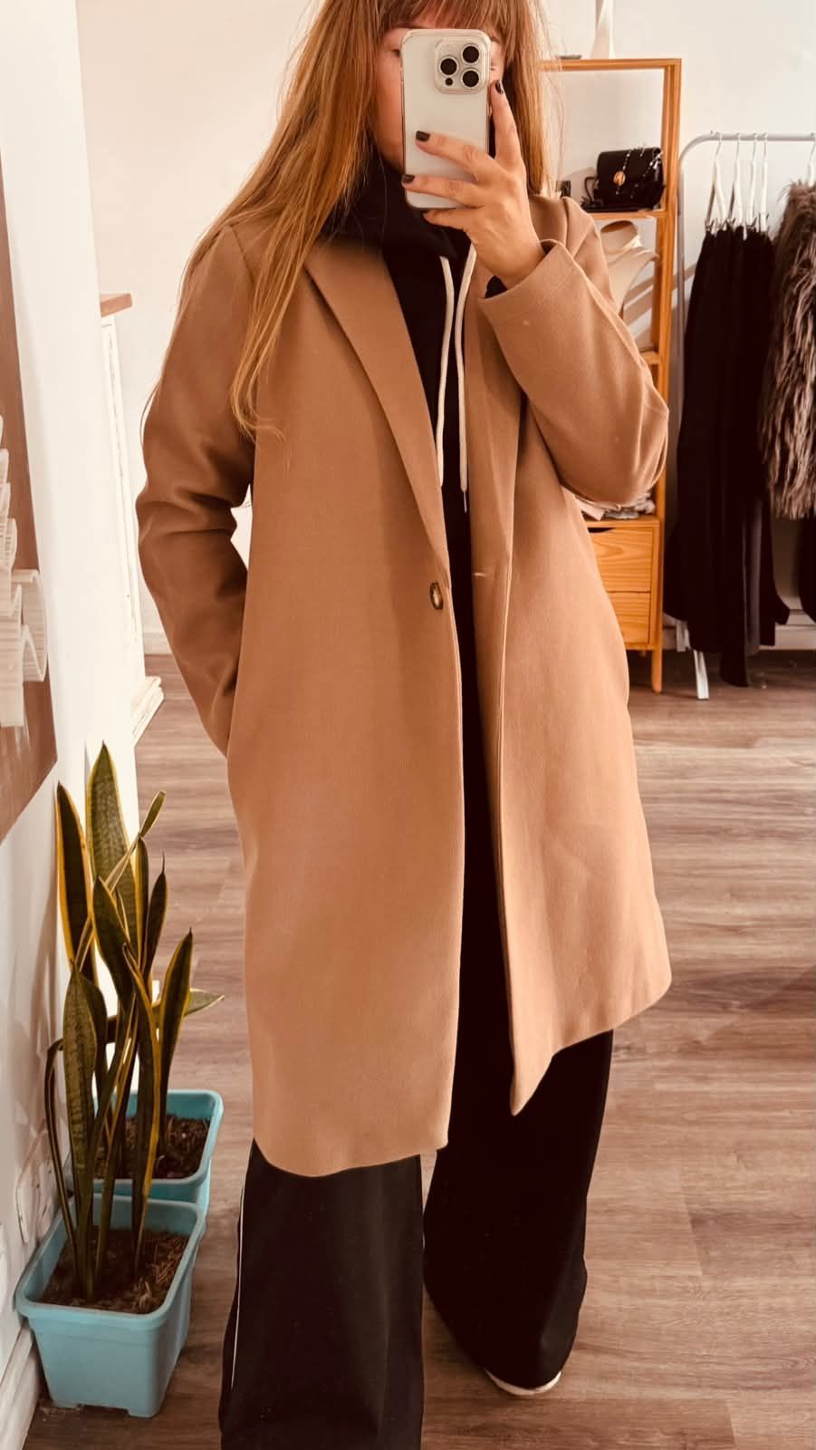 Una persona con un abrigo beige se toma una selfie frente al espejo en una tienda de ropa.
