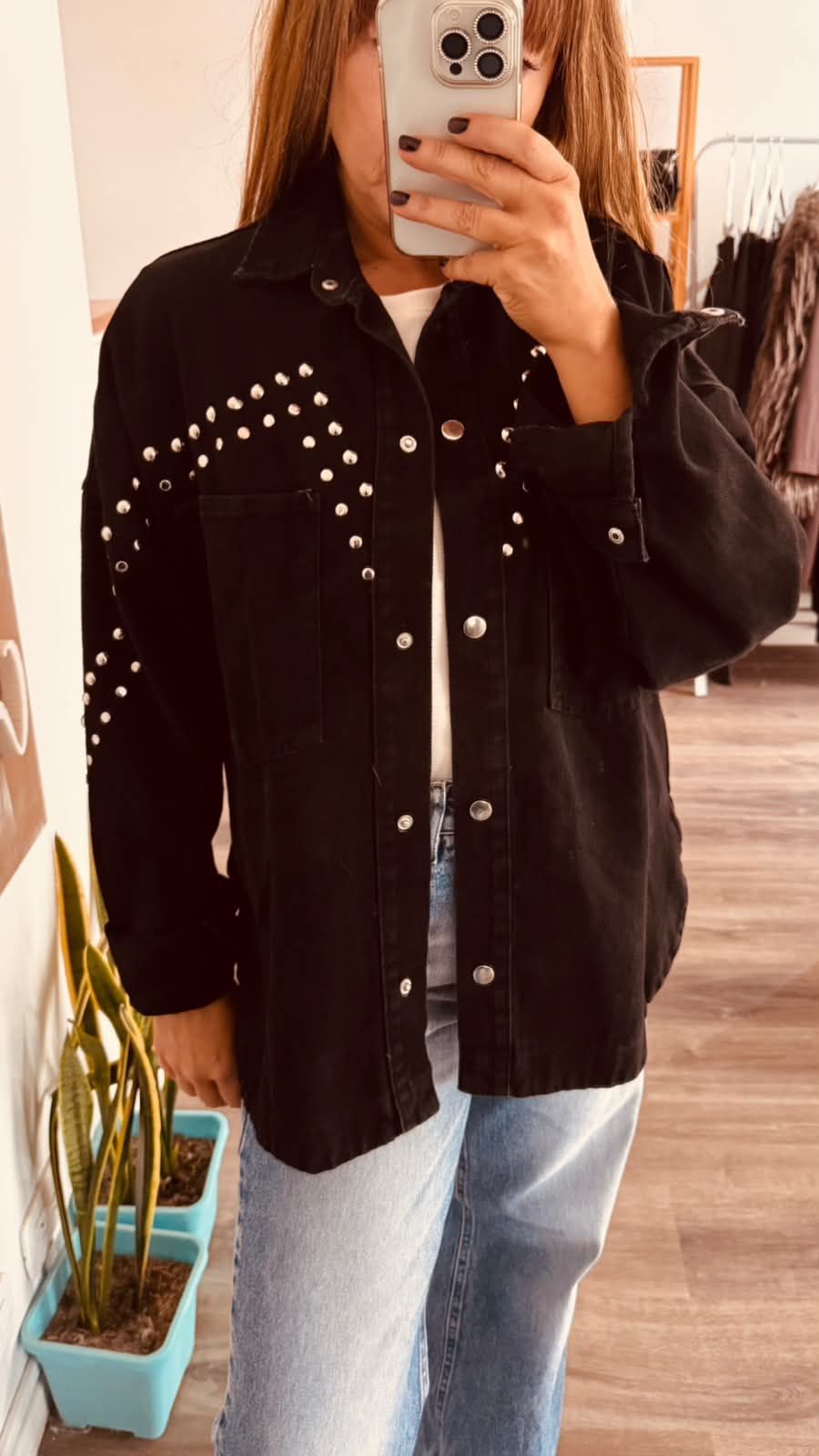 Persona tomándose una selfie frente al espejo con una camisa negra con tachuelas y jeans azul claro.