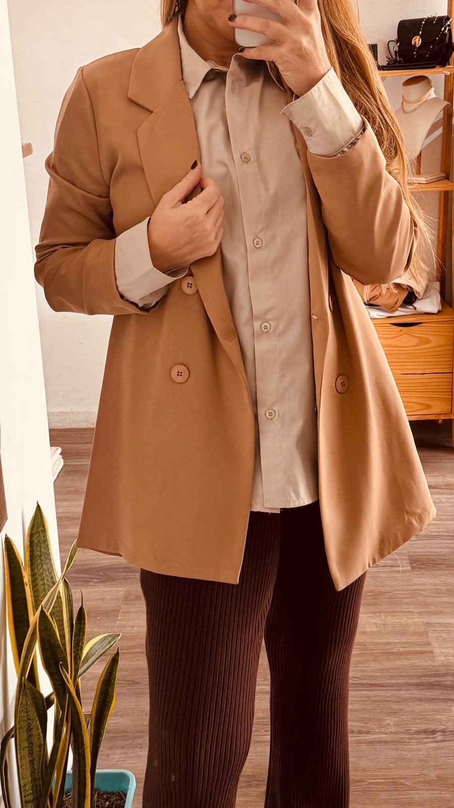 Persona tomándose una selfie frente al espejo con un abrigo color camel, una camisa beige y pantalones de rayas oscuras.