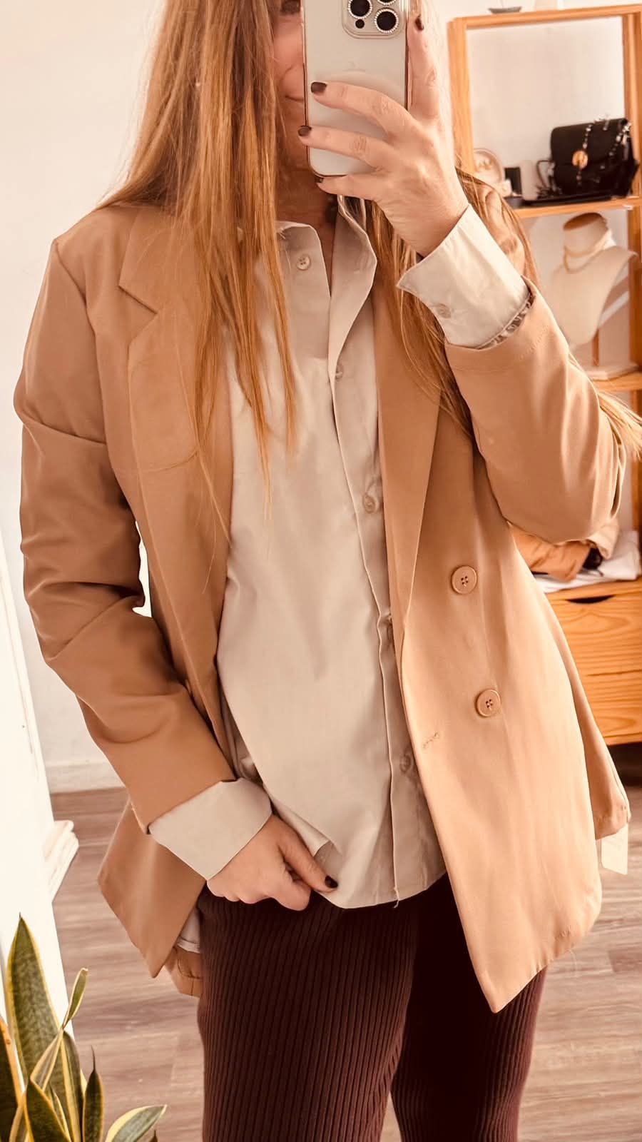 Persona tomándose una selfie frente al espejo con un abrigo color camel, una camisa beige y pantalones de rayas oscuras.