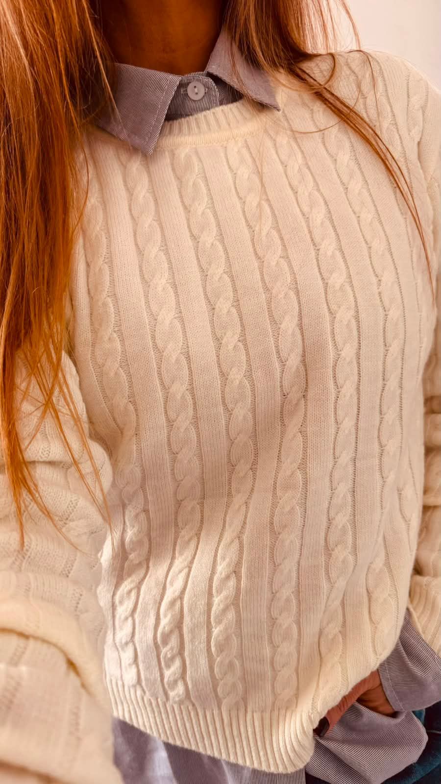 Una persona se toma una selfie frente al espejo con un suéter color crema sobre una camisa azul abotonada y jeans.