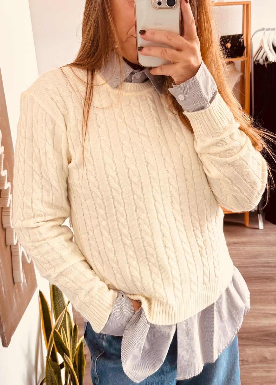 Una persona se toma una selfie frente al espejo con un suéter color crema sobre una camisa azul abotonada y jeans.