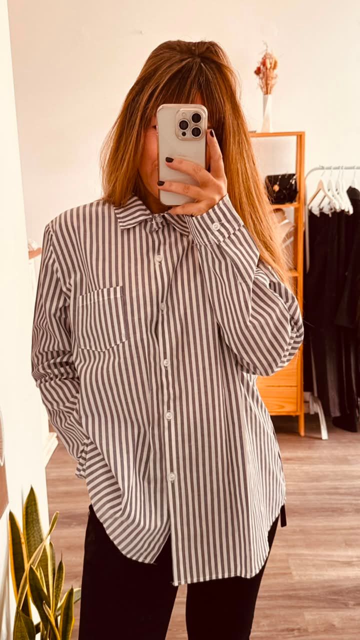 Una persona se toma una selfie frente al espejo, vestida con una camisa de rayas extragrande, en una habitación luminosa.