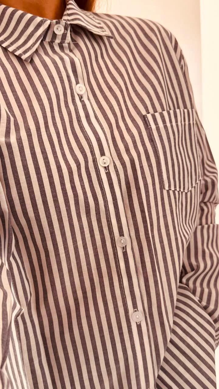 Una persona se toma una selfie frente al espejo, vestida con una camisa de rayas extragrande, en una habitación luminosa.