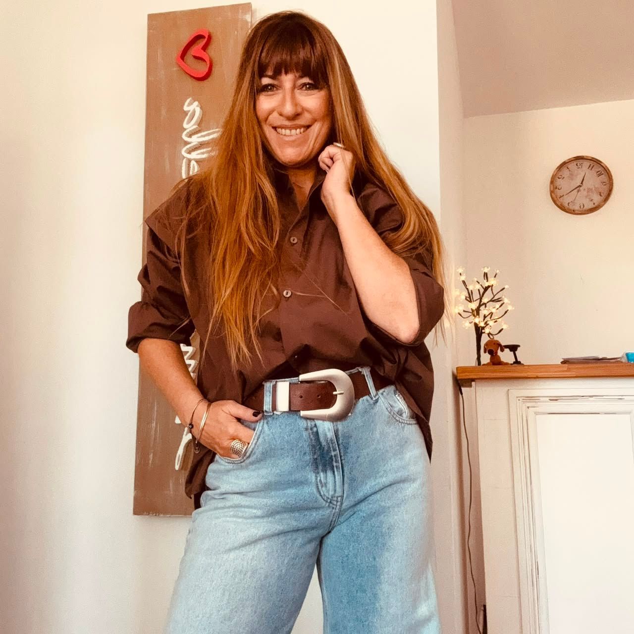 Mujer sonriente con camisa marrón y vaqueros de pie en el interior de una casa junto a un armario y un cuadro.