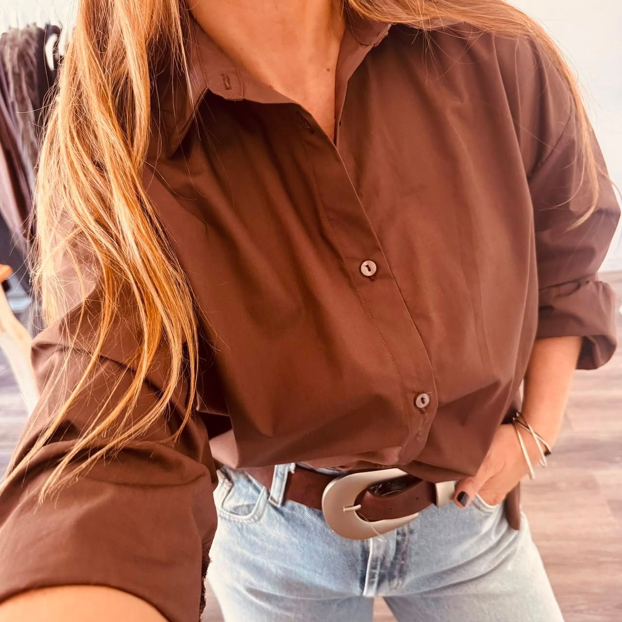 Mujer sonriente con camisa marrón y vaqueros de pie en el interior de una casa junto a un armario y un cuadro.