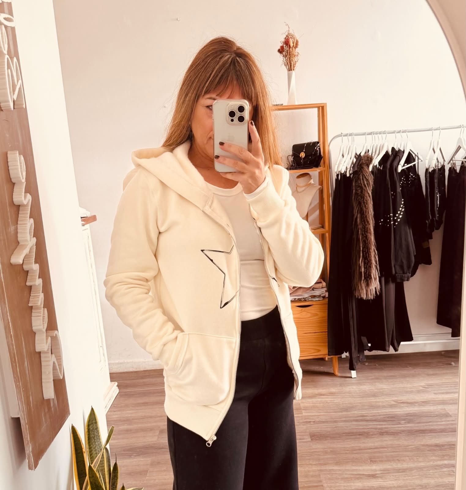 Una persona se toma una selfie frente al espejo con una sudadera con capucha color crema y pantalones negros en un vestidor.