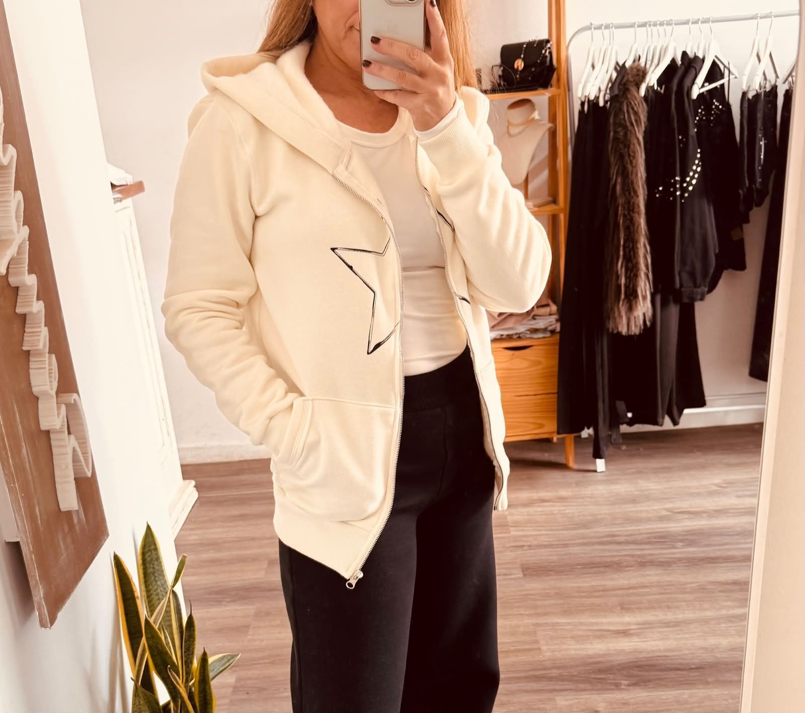 Una persona se toma una selfie frente al espejo con una sudadera con capucha color crema y pantalones negros en un vestidor.