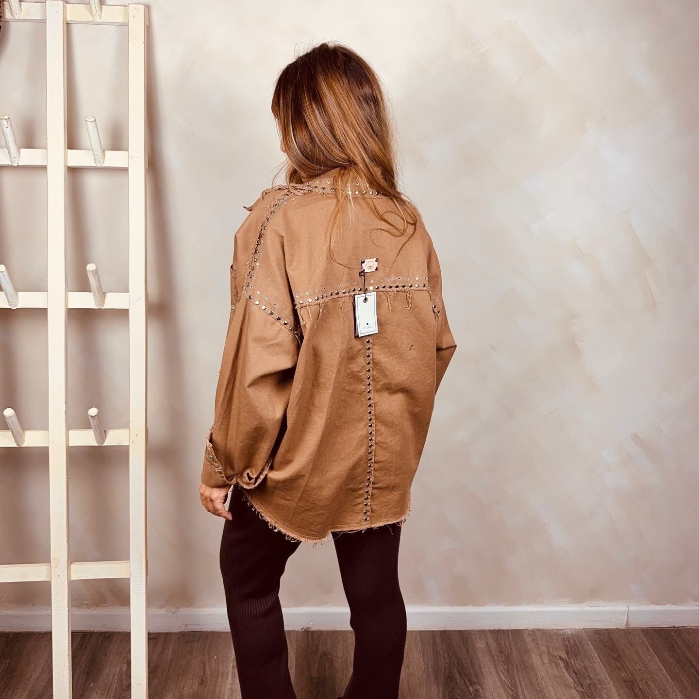 Mujer de pie en el interior de un edificio, vestida con una chaqueta color canela, una blusa blanca y pantalones oscuros.