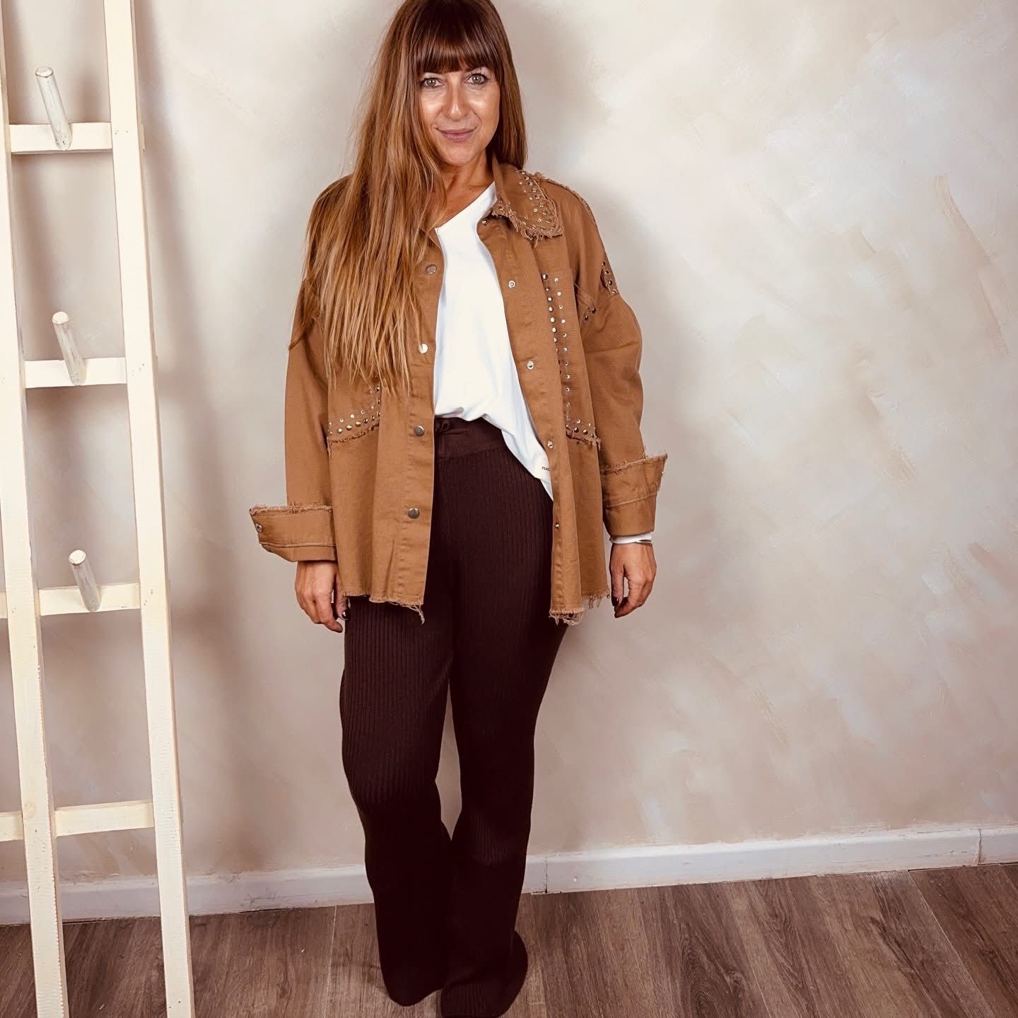 Mujer de pie en el interior de un edificio, vestida con una chaqueta color canela, una blusa blanca y pantalones oscuros.