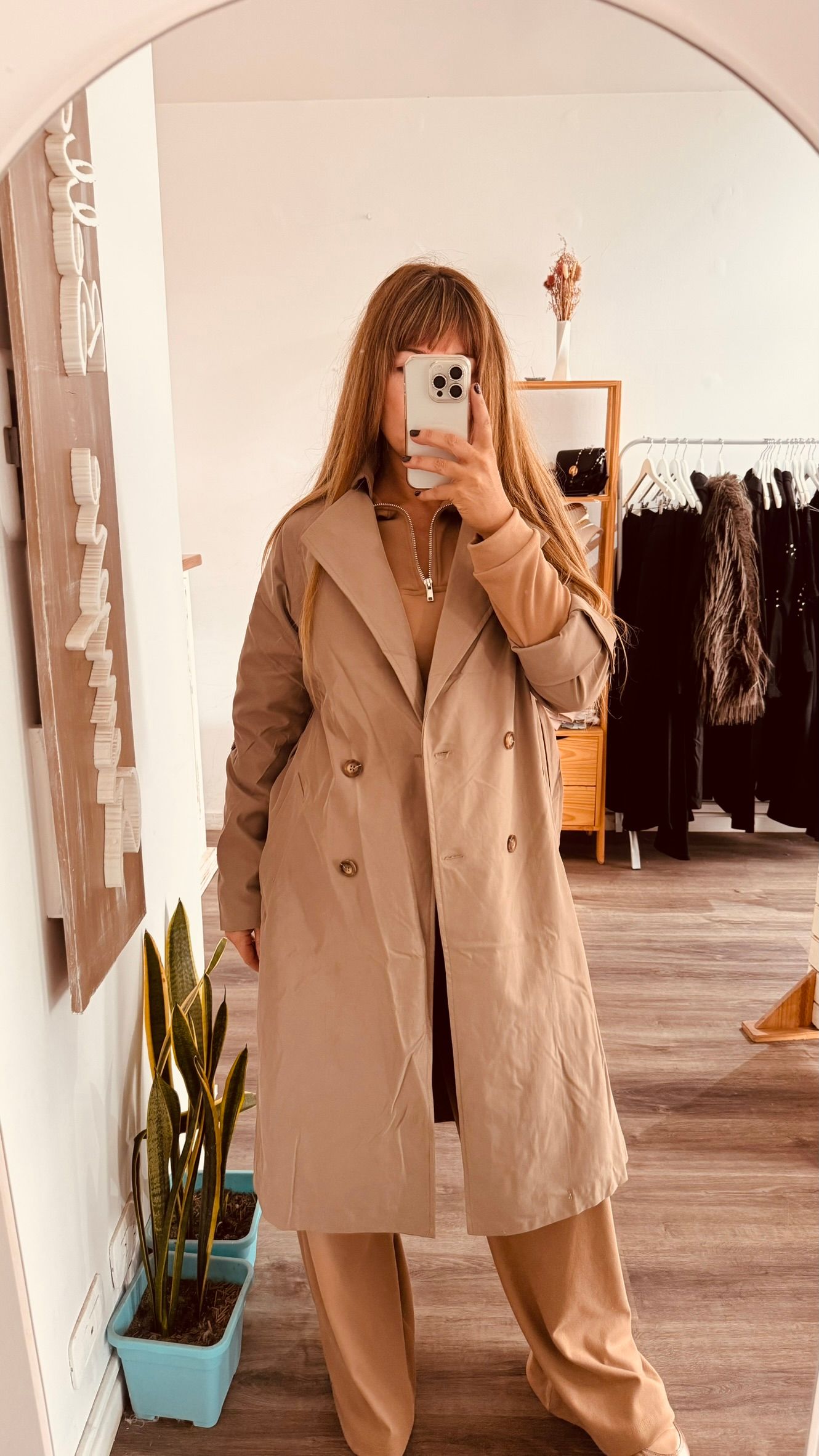 Una persona con una gabardina beige se toma una selfie frente al espejo en una tienda de ropa.