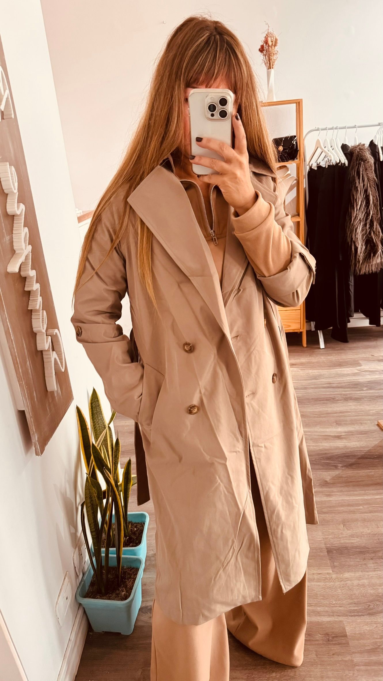 Una persona con una gabardina beige se toma una selfie frente al espejo en una tienda de ropa.