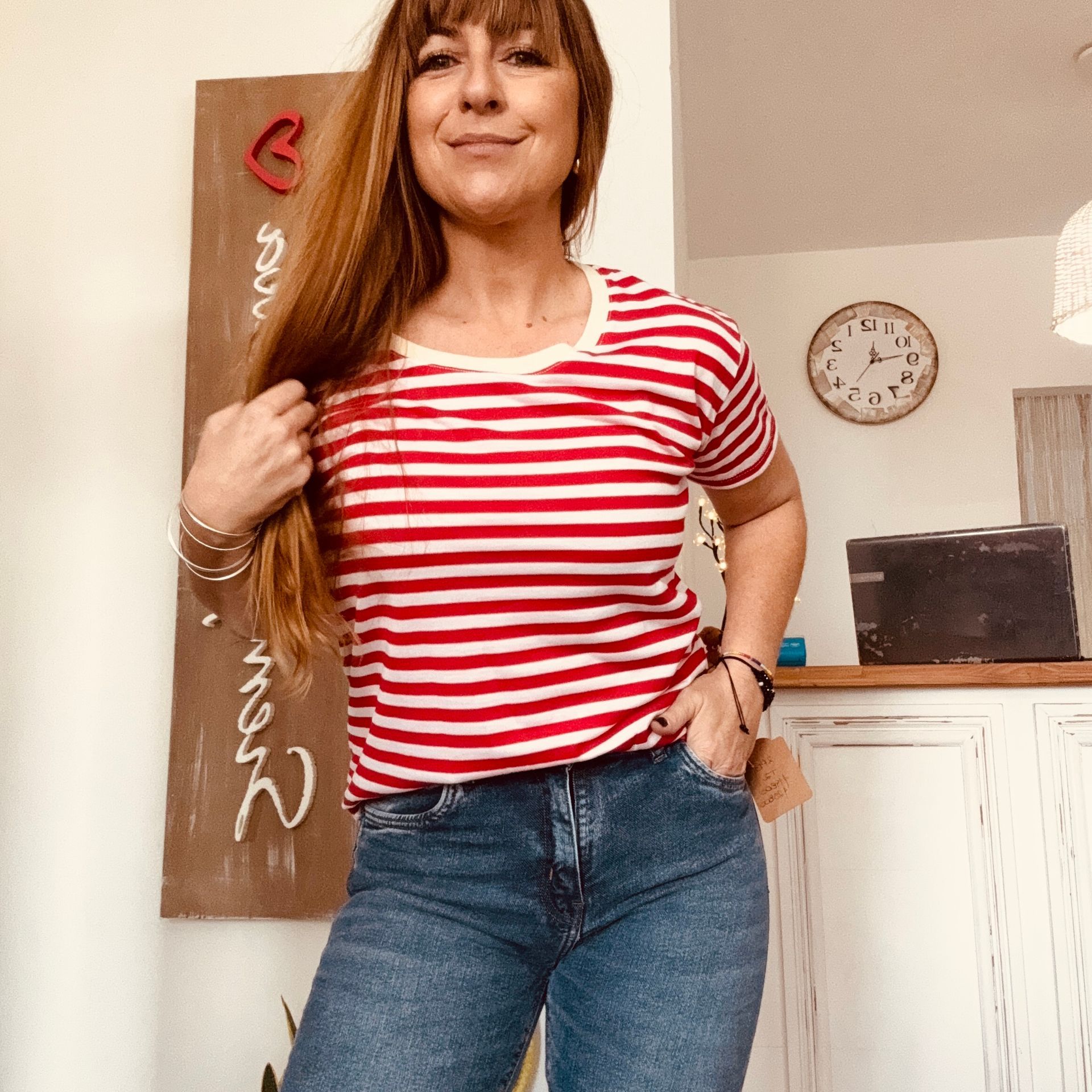 Mujer con camisa de rayas rojas y blancas y vaqueros, de pie en el interior de un edificio, sonriendo con las manos en los bolsillos.