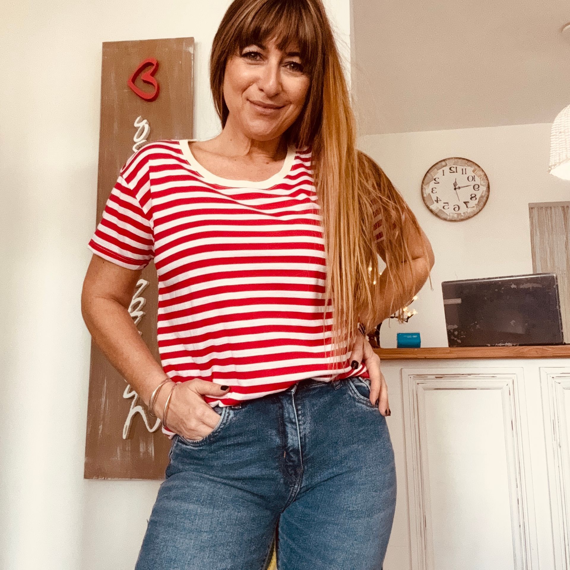 Mujer con camisa de rayas rojas y blancas y vaqueros, de pie en el interior de un edificio, sonriendo con las manos en los bolsillos.