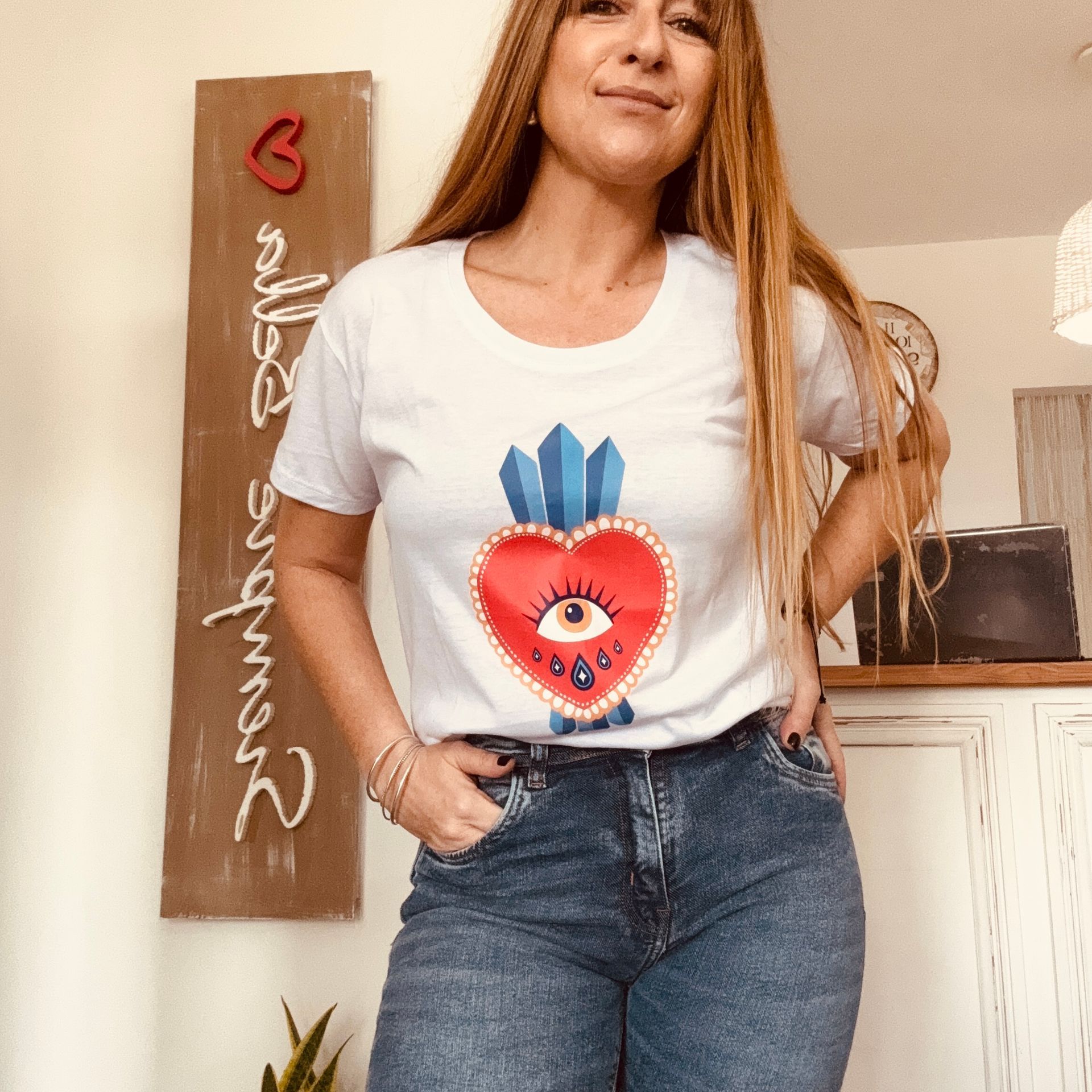 Mujer de pie en el interior de una habitación luminosa, vestida con una camiseta blanca estampada y vaqueros, con las manos en los bolsillos.