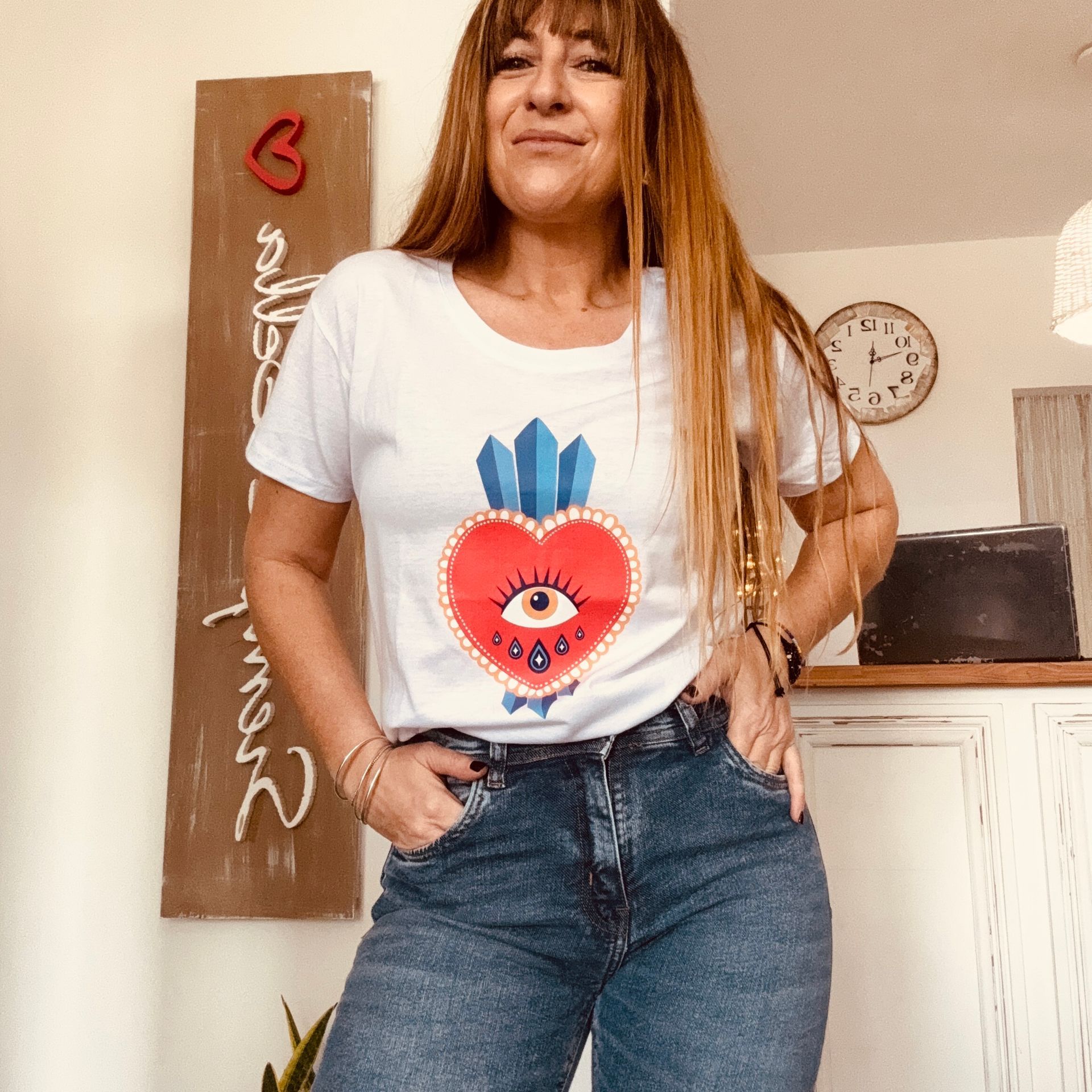 Mujer de pie en el interior de una habitación luminosa, vestida con una camiseta blanca estampada y vaqueros, con las manos en los bolsillos.