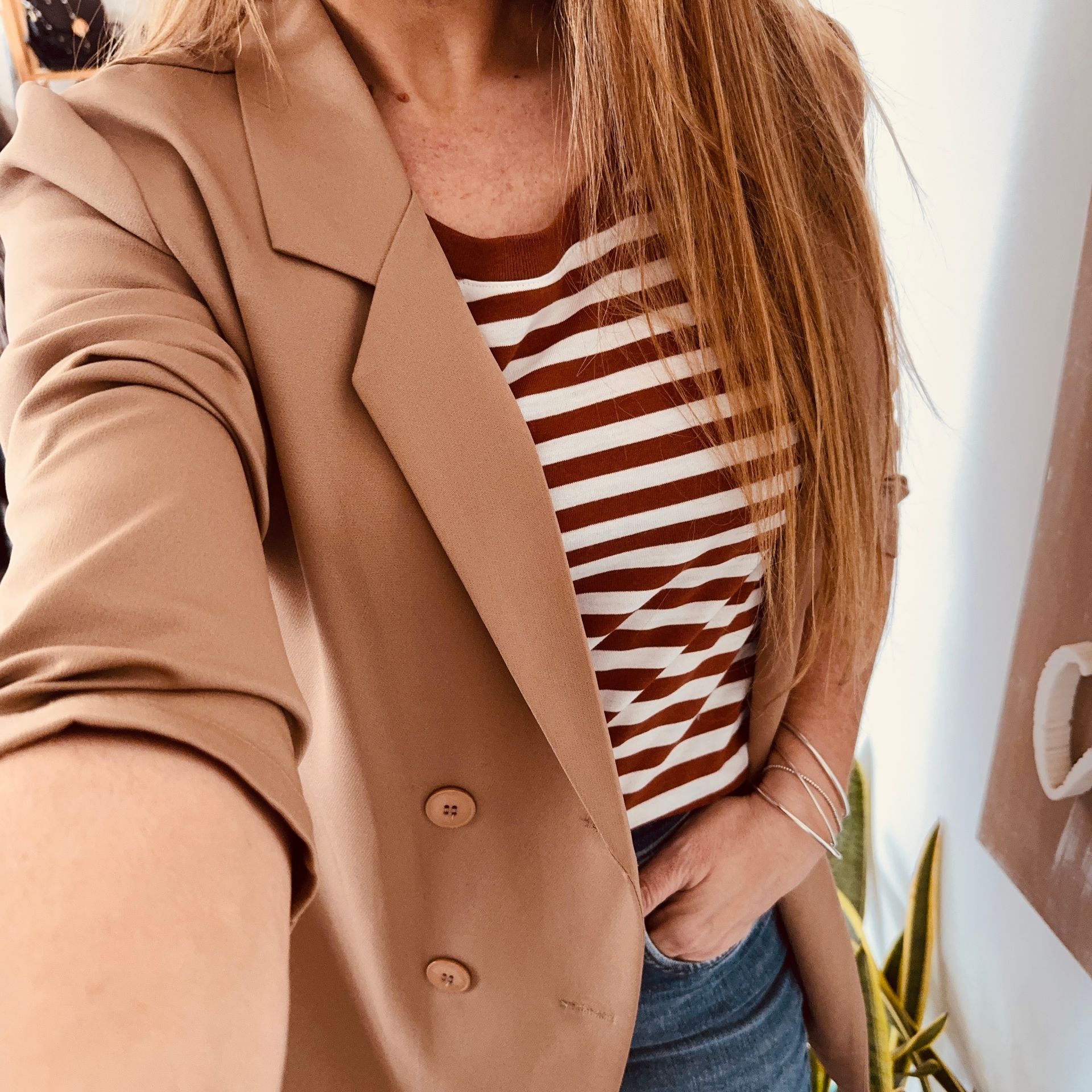 Mujer con abrigo color canela y blusa de rayas posando en el interior junto a una pared con un reloj y elementos decorativos.