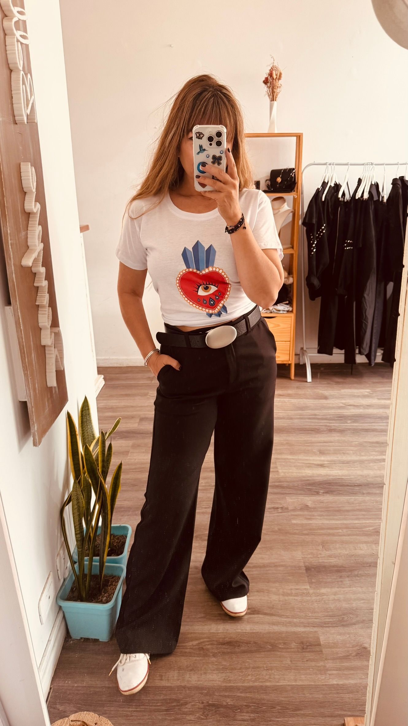 Una persona se toma una selfie frente al espejo en una tienda de ropa, vistiendo una camiseta blanca estampada y jeans oscuros.