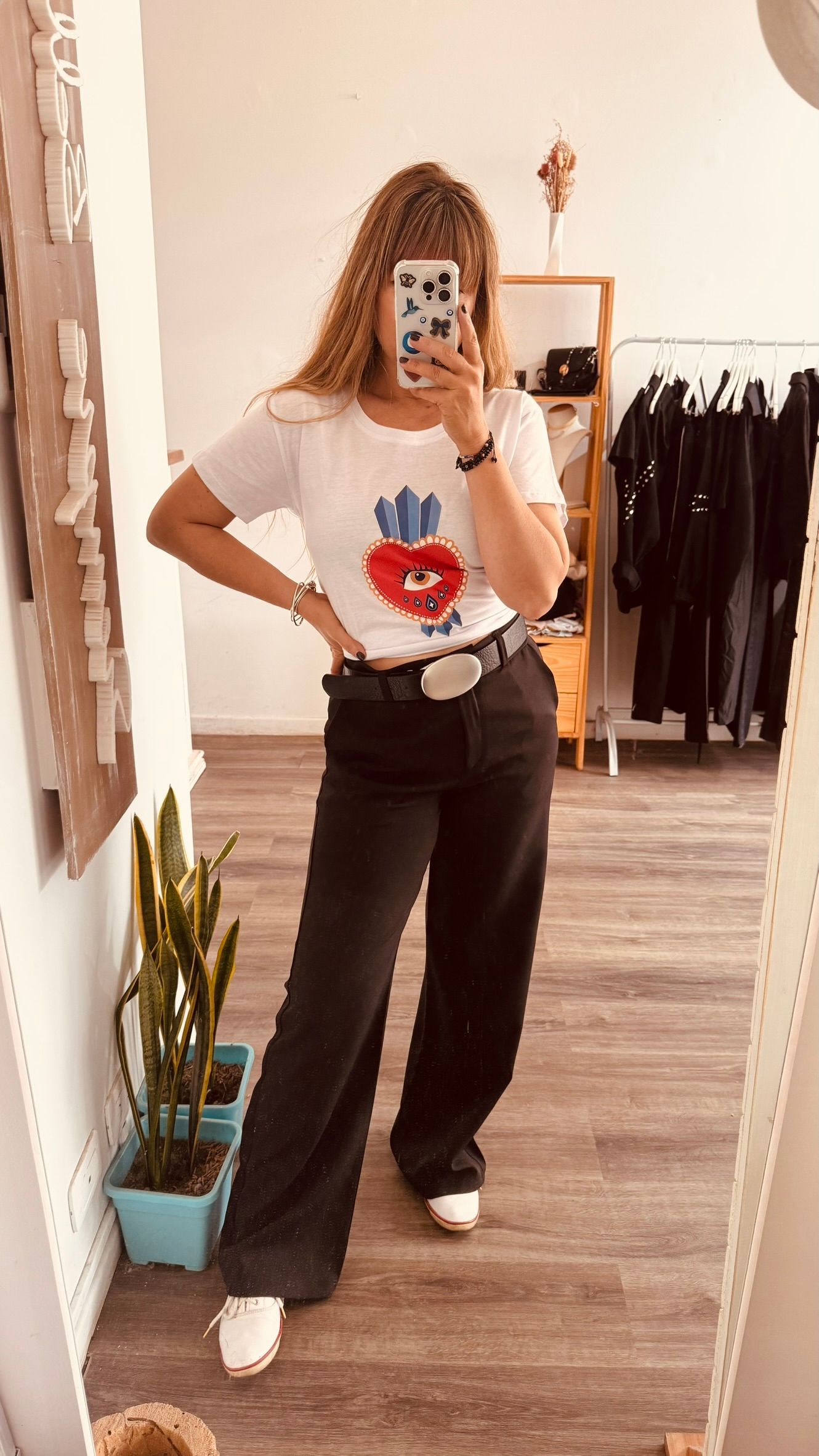 Una persona se toma una selfie frente al espejo en una tienda de ropa, vistiendo una camiseta blanca estampada y jeans oscuros.