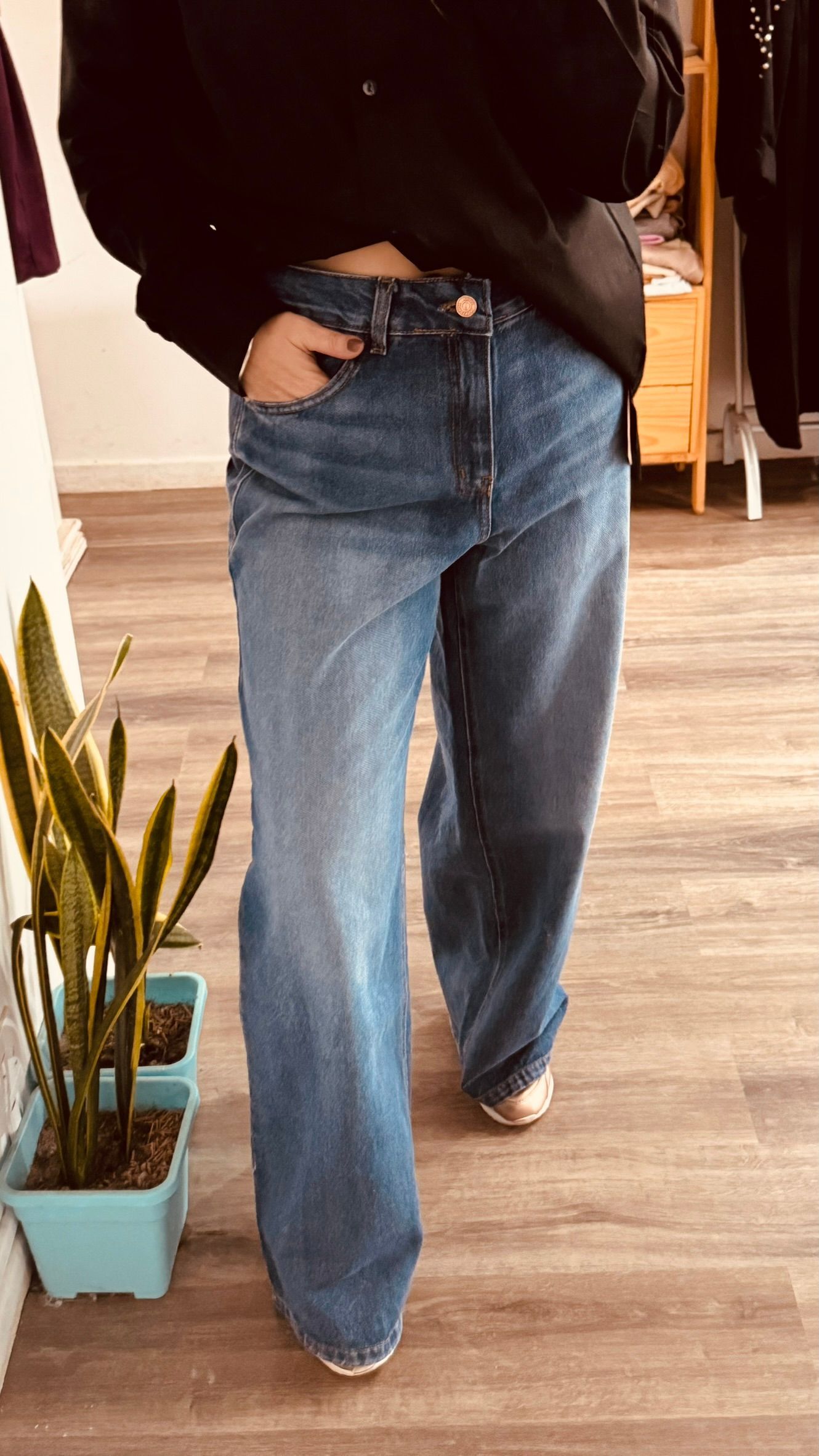 Persona de pie en el interior de un edificio, vestida con una camiseta negra y pantalones vaqueros azules, con una mano en el bolsillo.