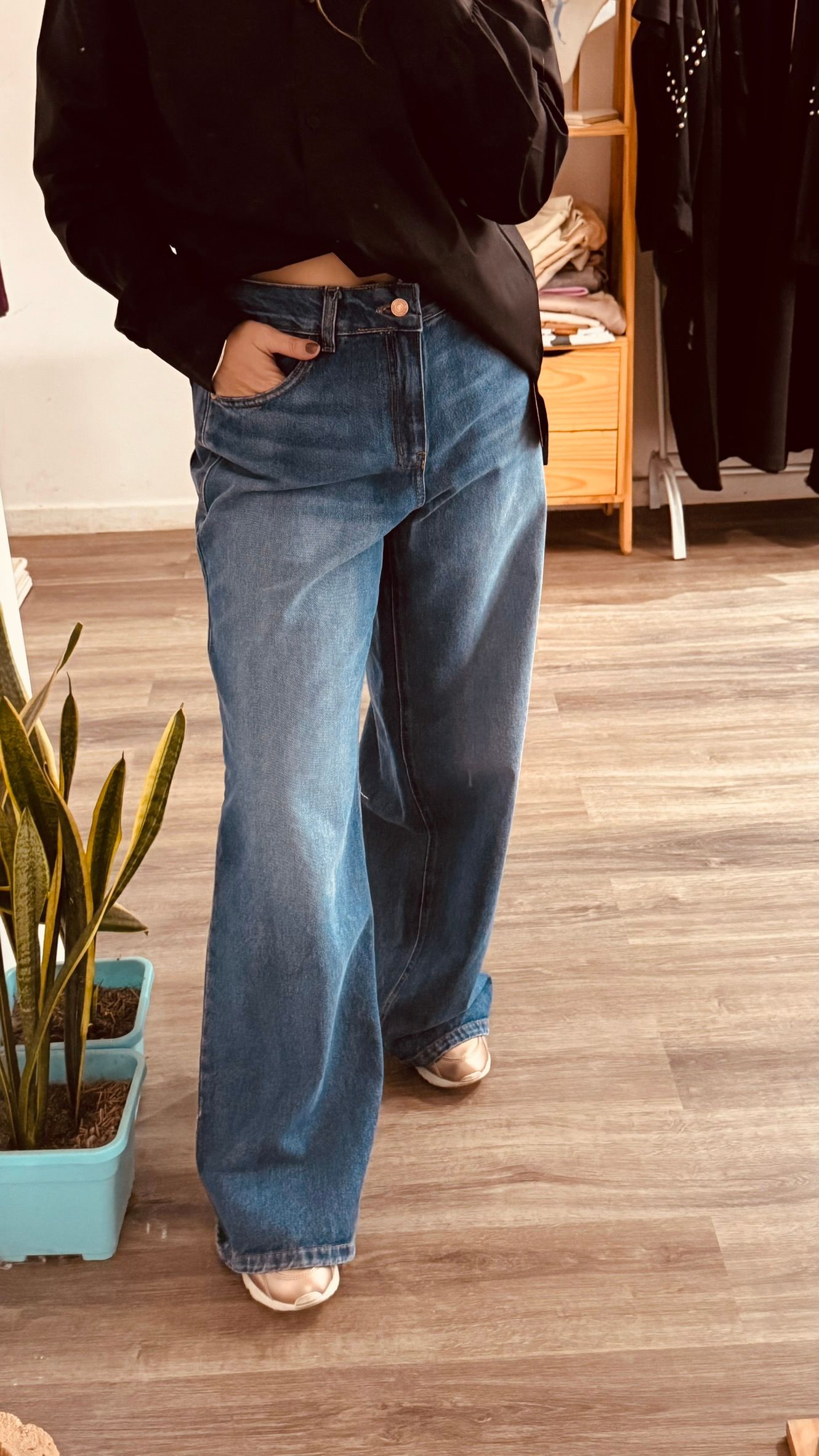 Persona de pie en el interior de un edificio, vestida con una camiseta negra y pantalones vaqueros azules, con una mano en el bolsillo.