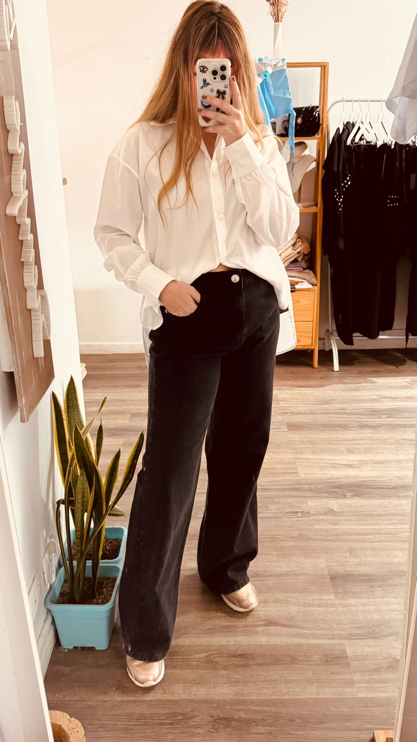 Una persona se toma una selfie frente al espejo en una tienda de ropa, vestida con una blusa blanca, pantalones negros y zapatillas deportivas.
