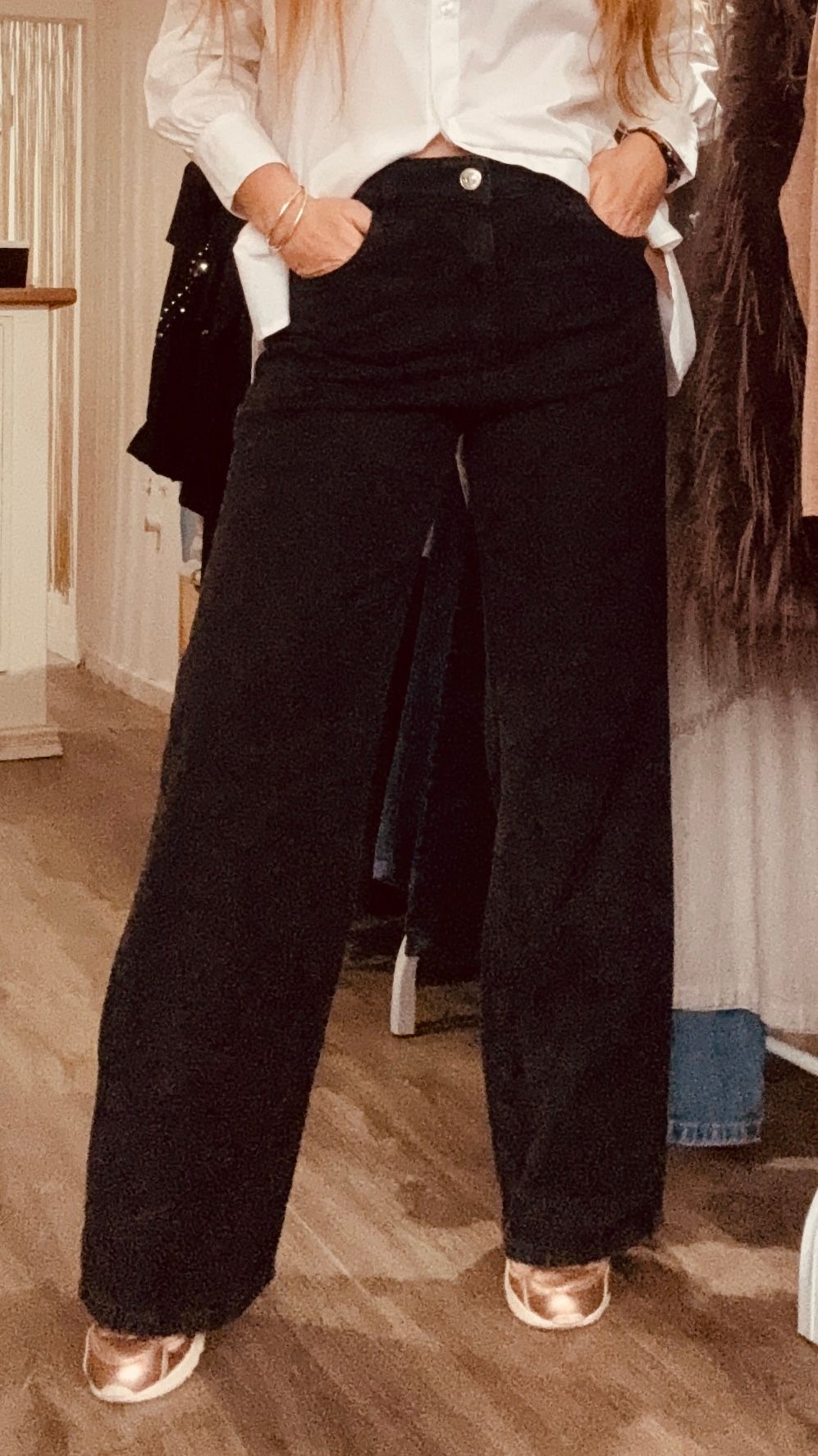Una persona se toma una selfie frente al espejo en una tienda de ropa, vestida con una blusa blanca, pantalones negros y zapatillas deportivas.