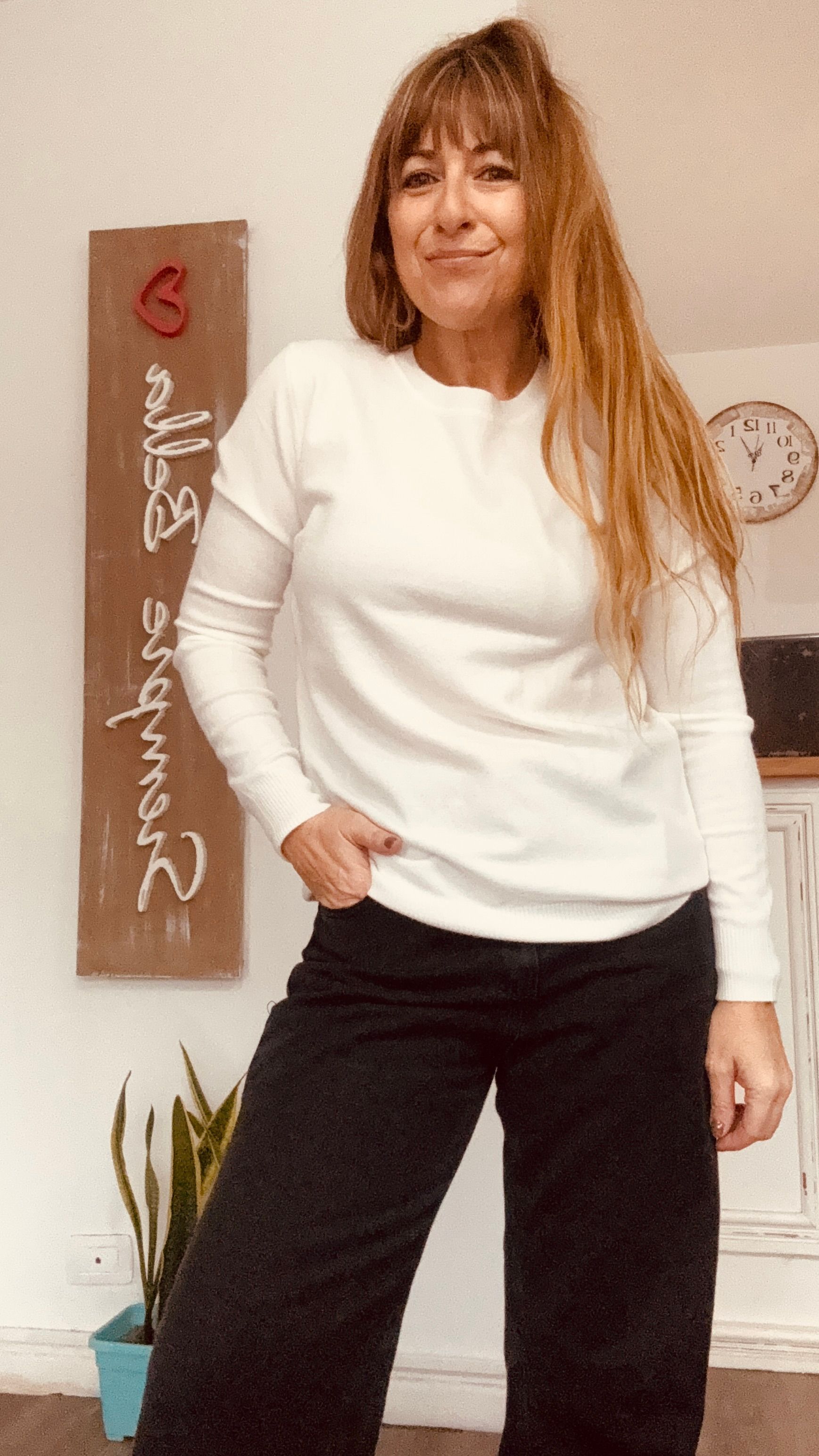 Mujer con suéter blanco y pantalones negros posando en el interior junto a un elemento decorativo de pared y una planta.