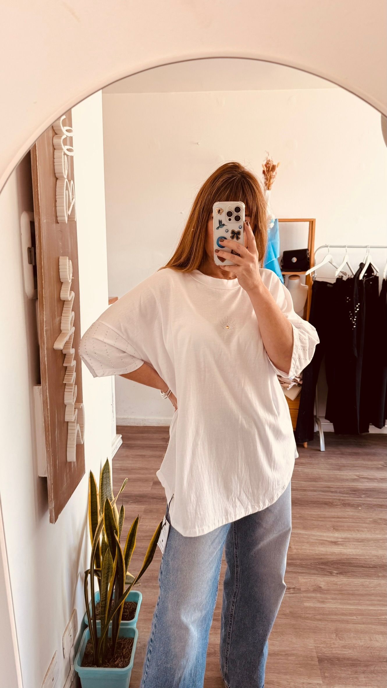 Una persona se toma una selfie frente al espejo, vestida con una blusa blanca y jeans azules, de pie en una habitación con un espejo arqueado.