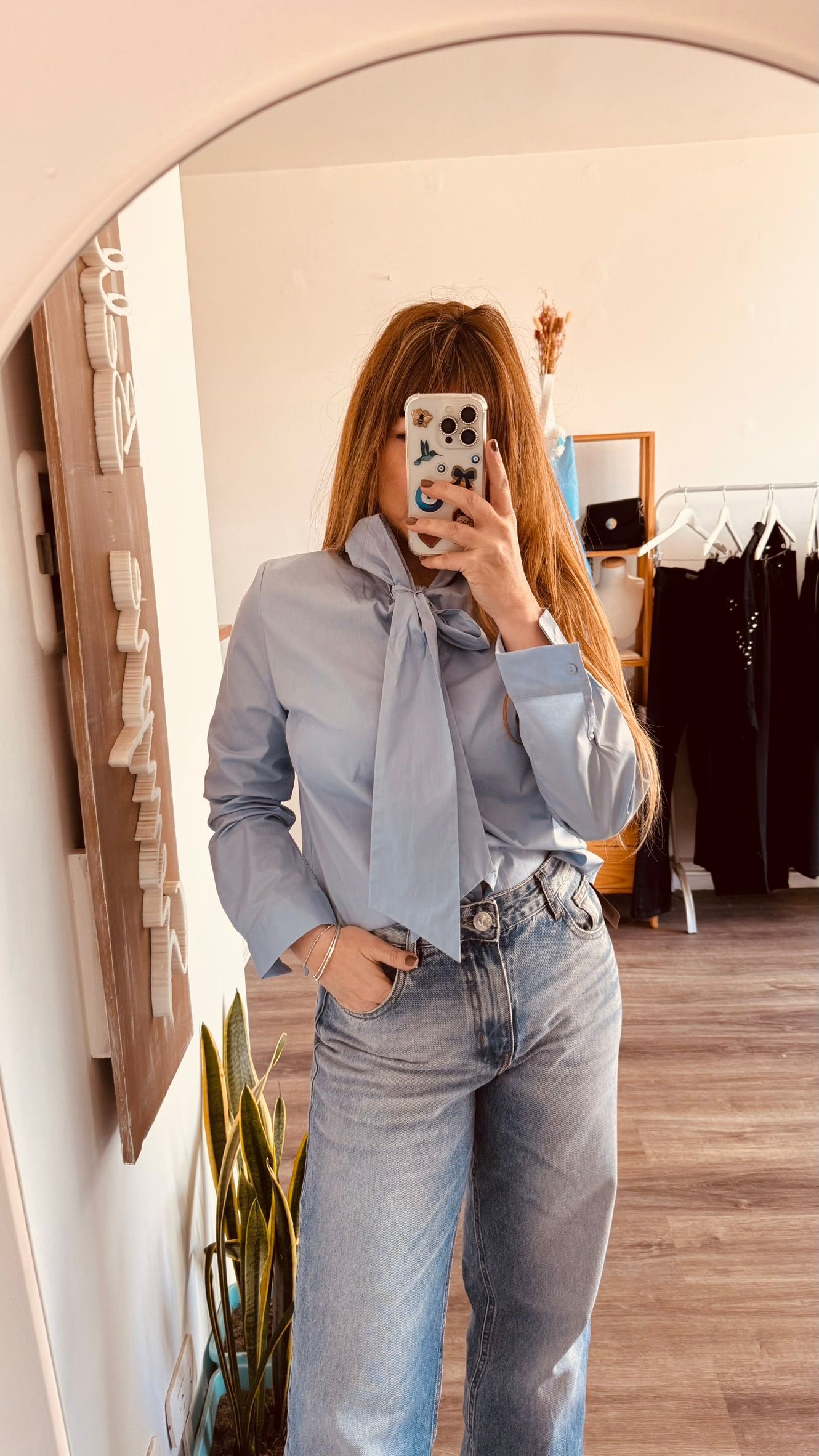 Una persona se toma una selfie frente al espejo, vestida con una camisa azul claro y jeans, de pie en un dormitorio.