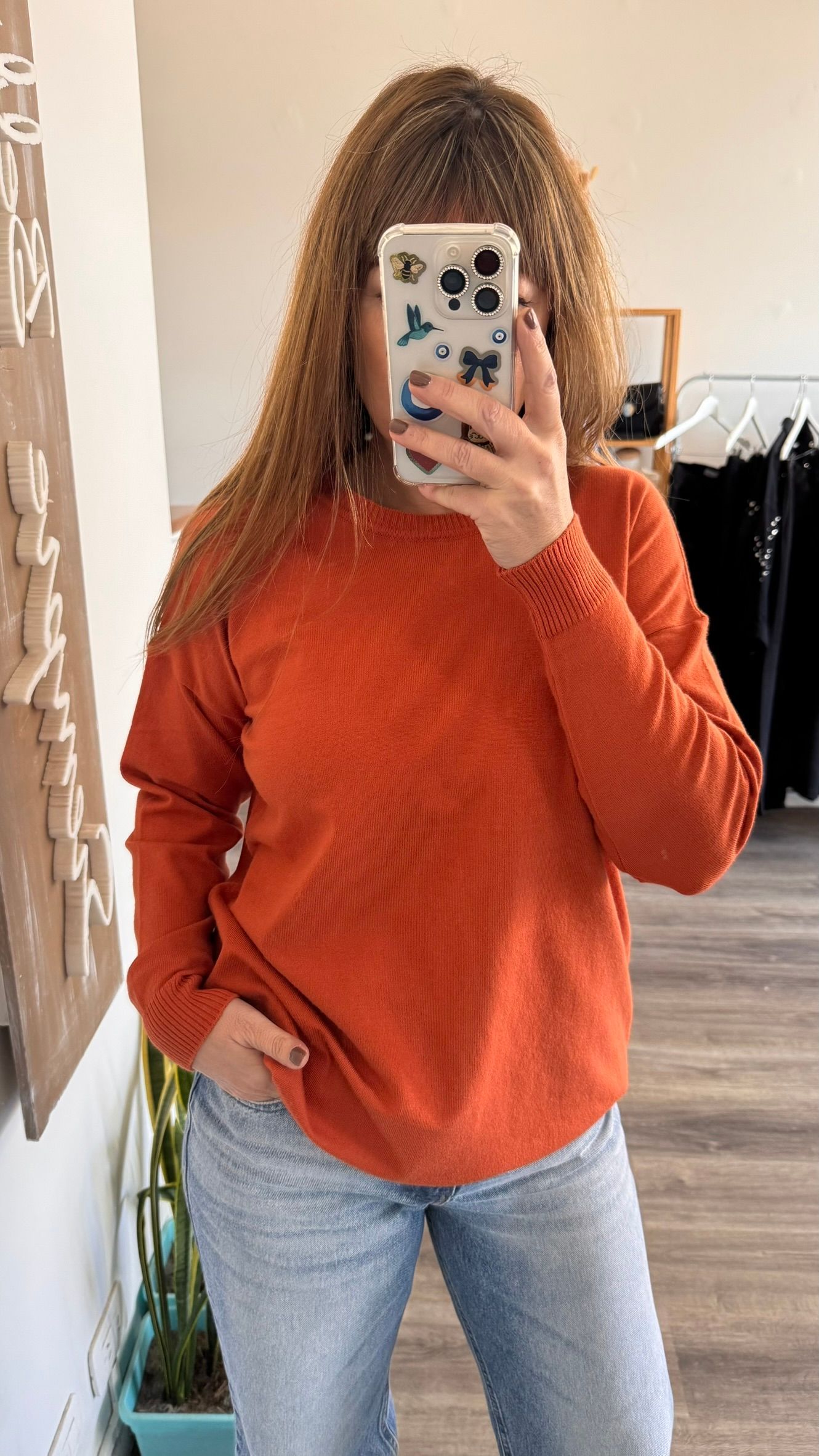 Una persona con un suéter naranja y jeans claros se toma una selfie frente a un espejo en el interior de una casa.