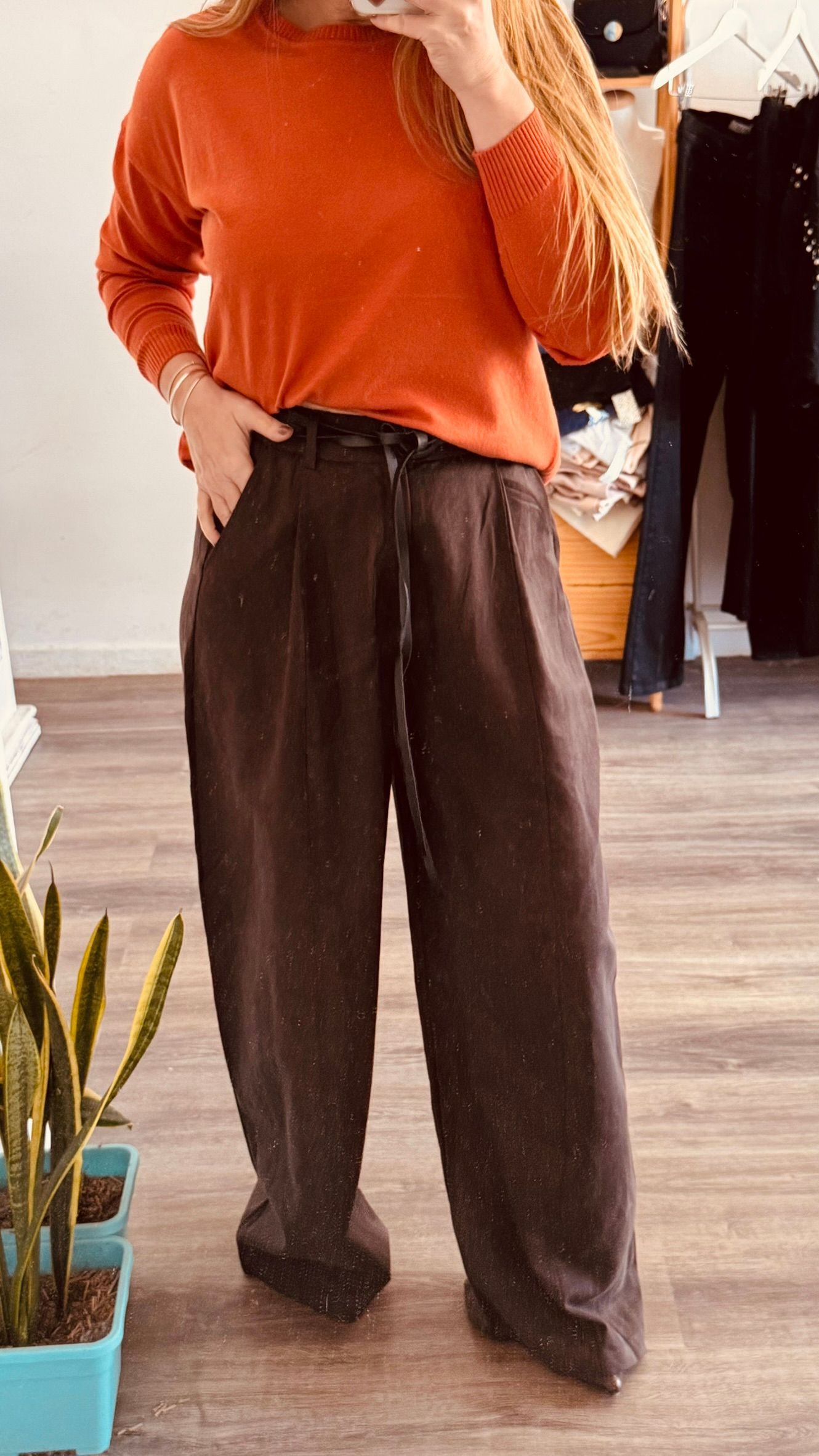 Persona con suéter naranja y pantalones anchos marrones de pie en el interior de un edificio.