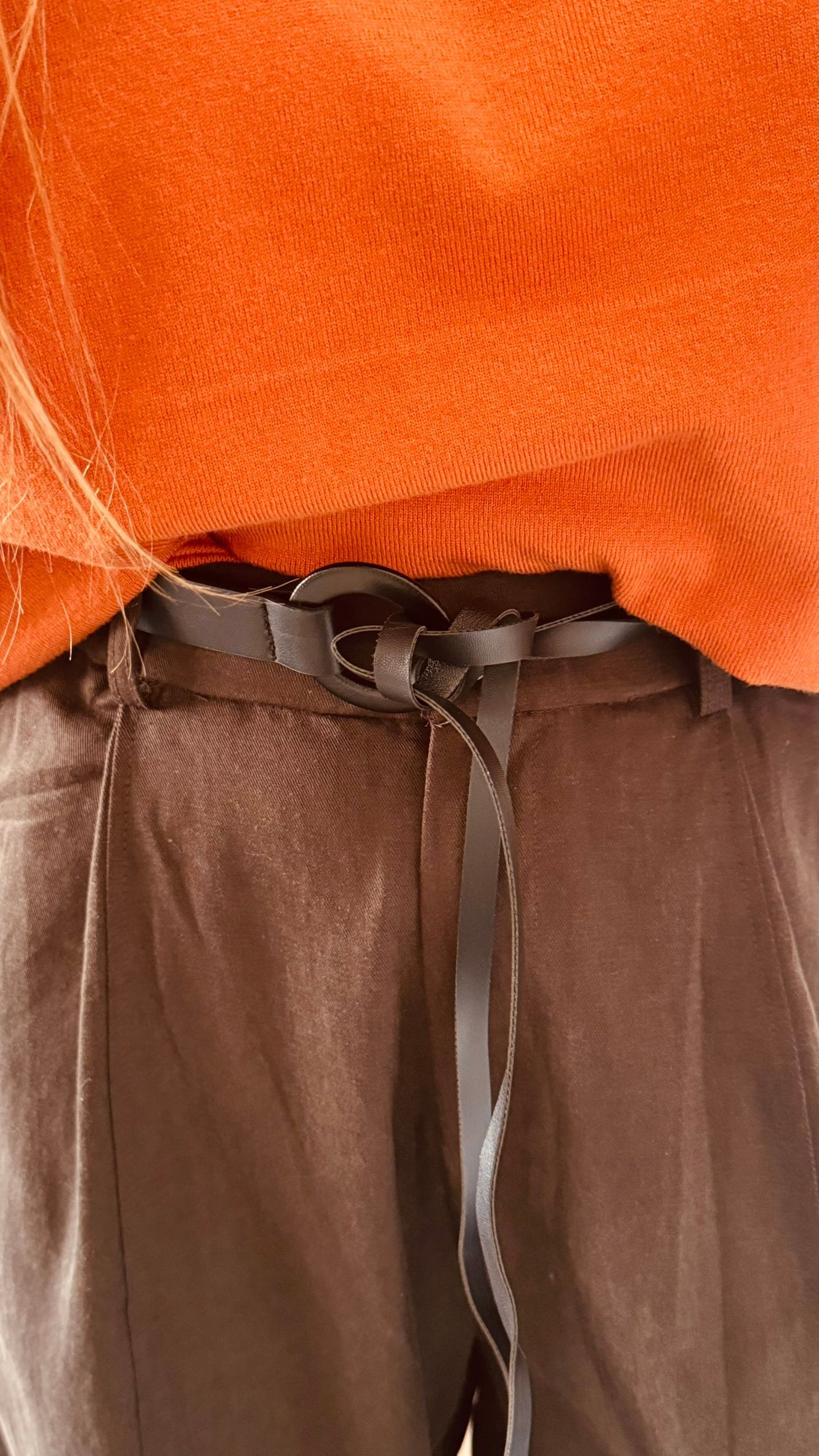Persona con suéter naranja y pantalones anchos marrones de pie en el interior de un edificio.