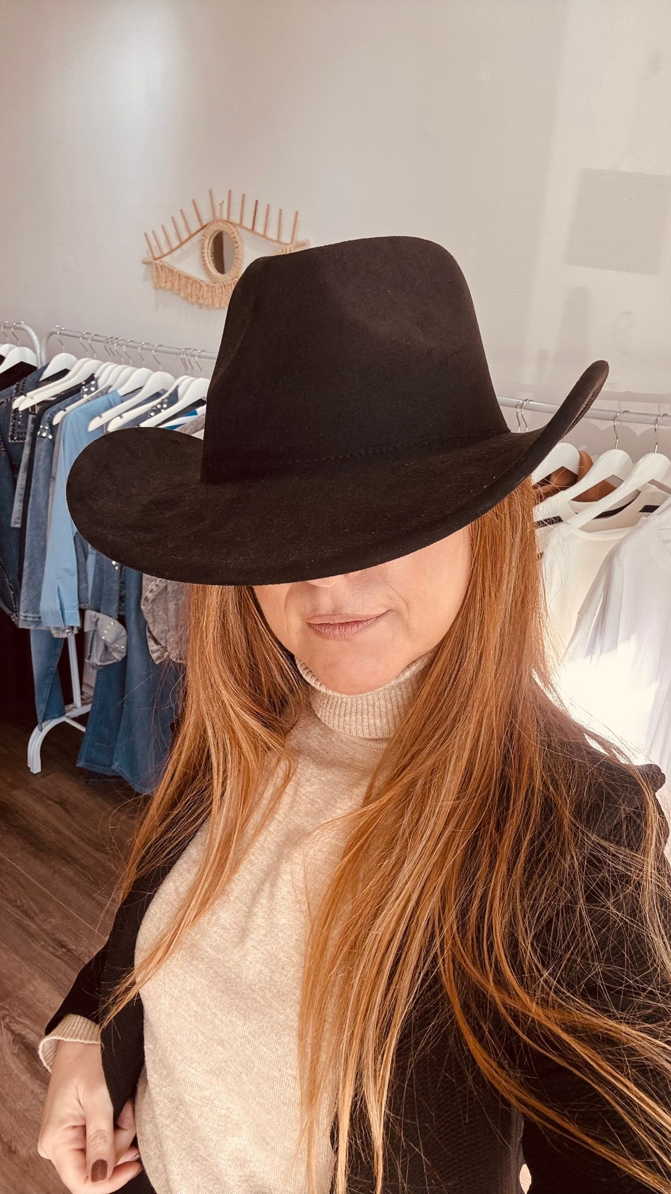 Mujer con sombrero y chaqueta de vaquero negros, posando en una tienda de ropa con perchas al fondo.