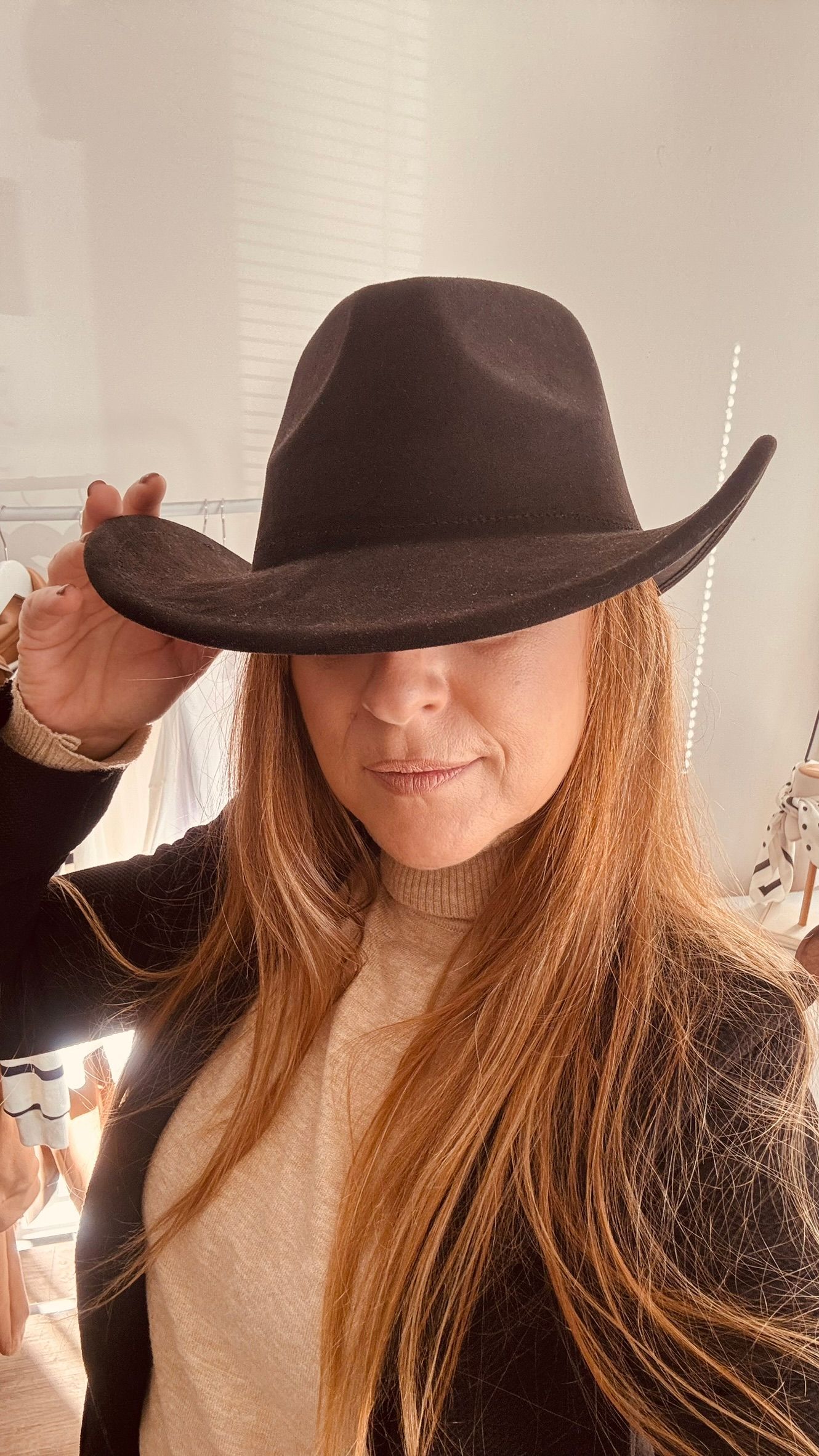 Mujer con sombrero y chaqueta de vaquero negros, posando en una tienda de ropa con perchas al fondo.