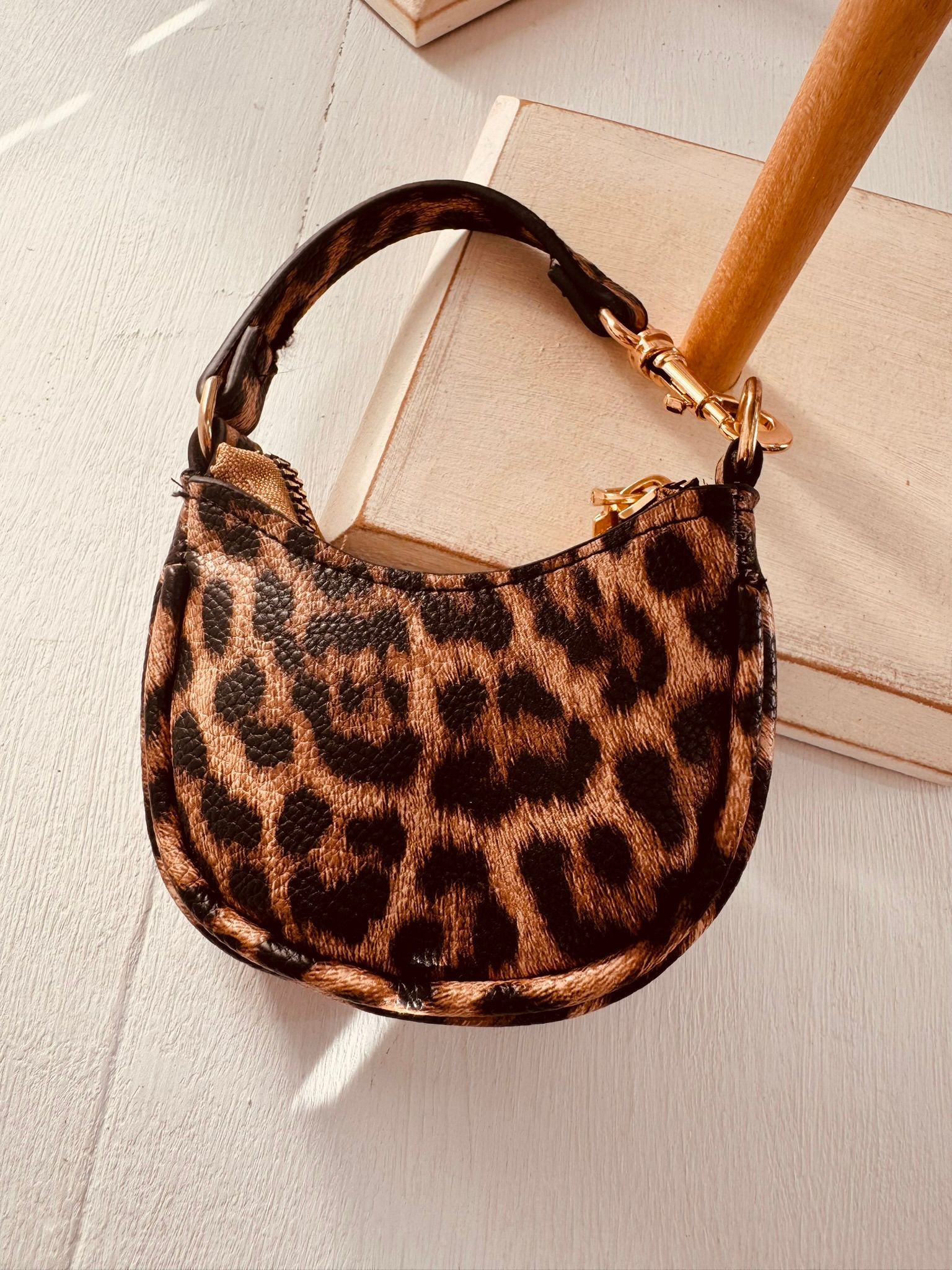 Una mano sostiene dos bolsos pequeños, uno negro y otro con estampado de leopardo, en el interior de una vivienda sobre un suelo de madera.